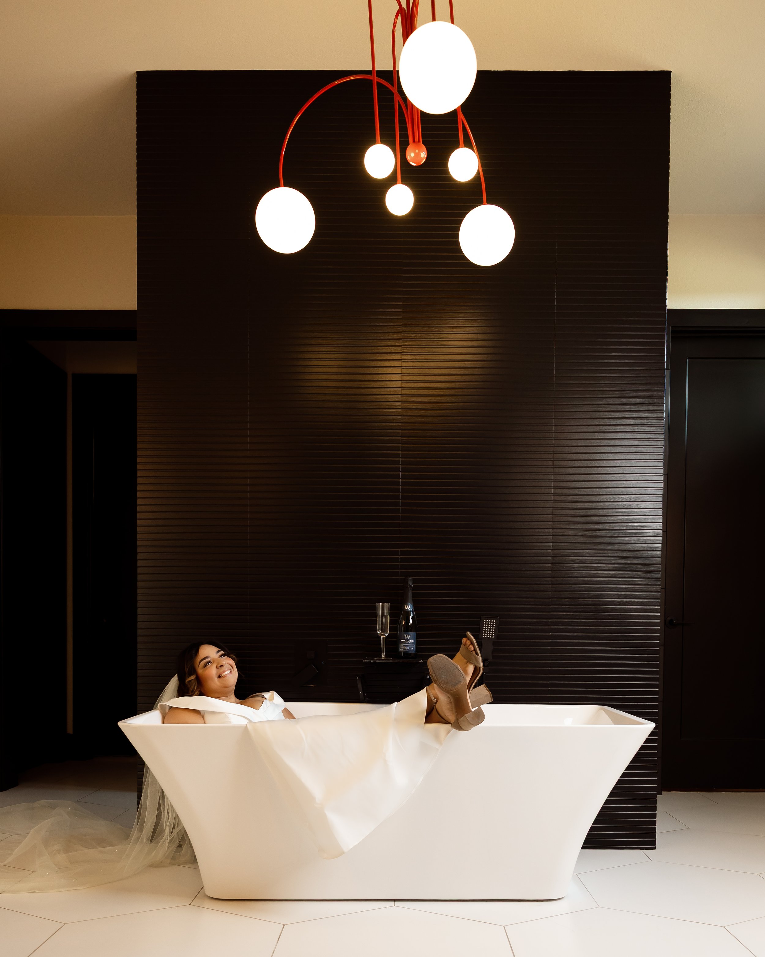 Woman relaxing in a white bathtub with her legs up, smiling, in a room with dark wood paneling and a black shelf holding a bottle of wine and a glass.