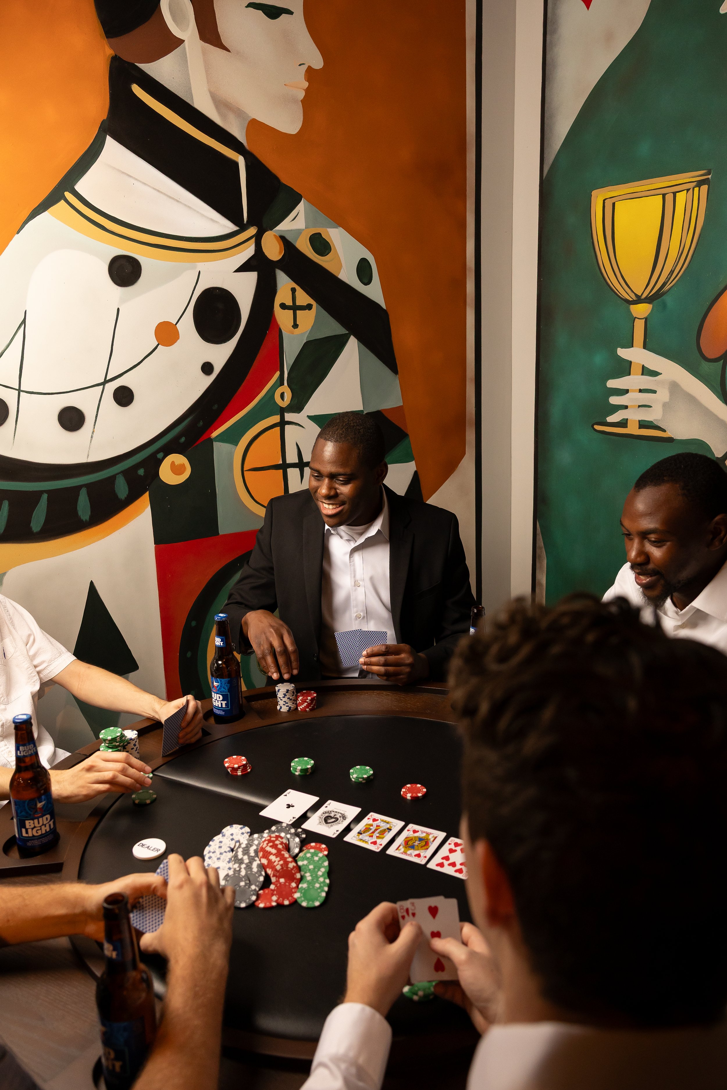 Group of people playing poker at a table with chips and drinks, in front of colorful abstract wall art.