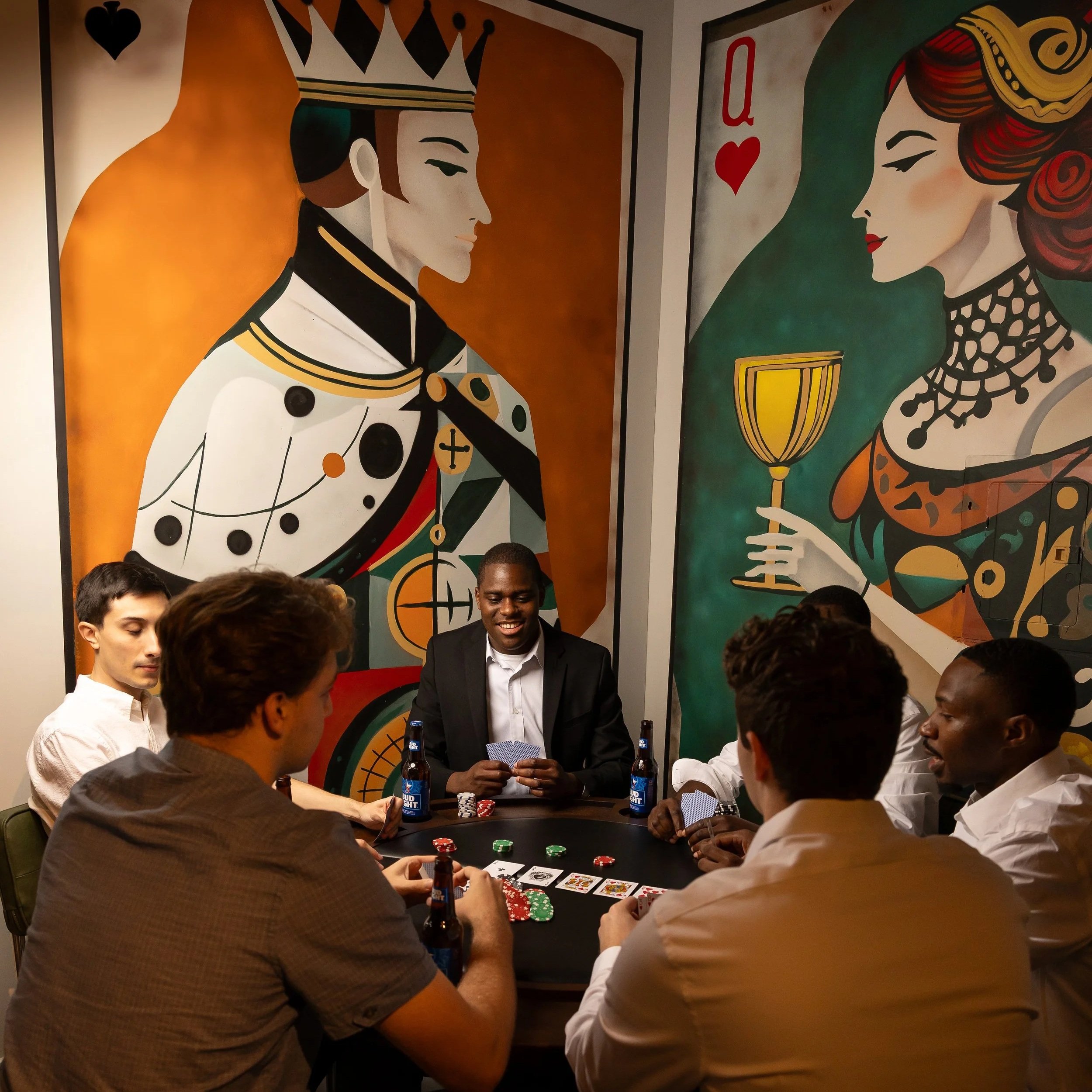 Six people playing poker at a table in a room with colorful, large playing card murals on the wall, depicting a king and a queen.