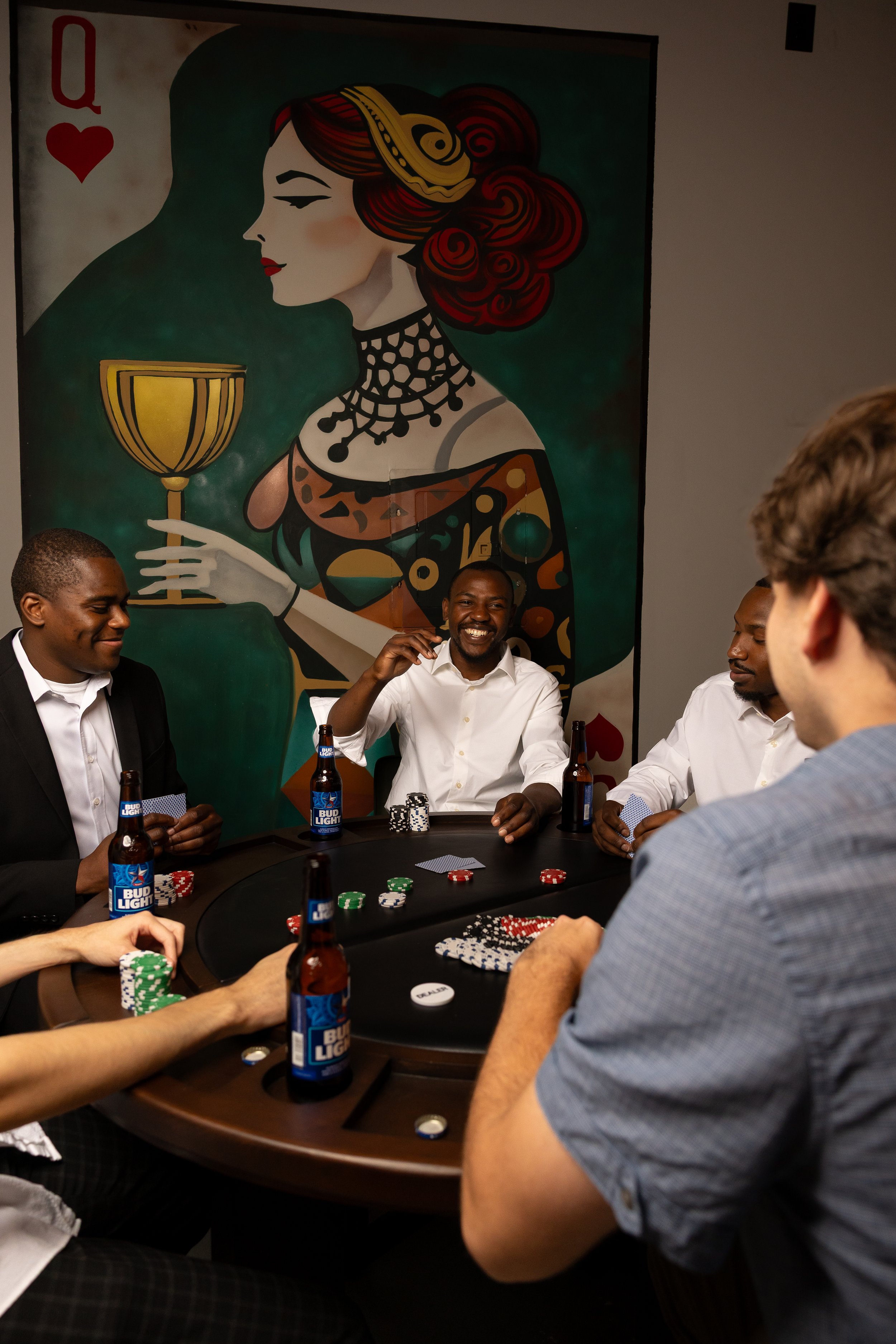 Women and men playing poker at a table in a game room with a large modern art portrait of a woman with red hair holding a glass of wine on the wall behind them.