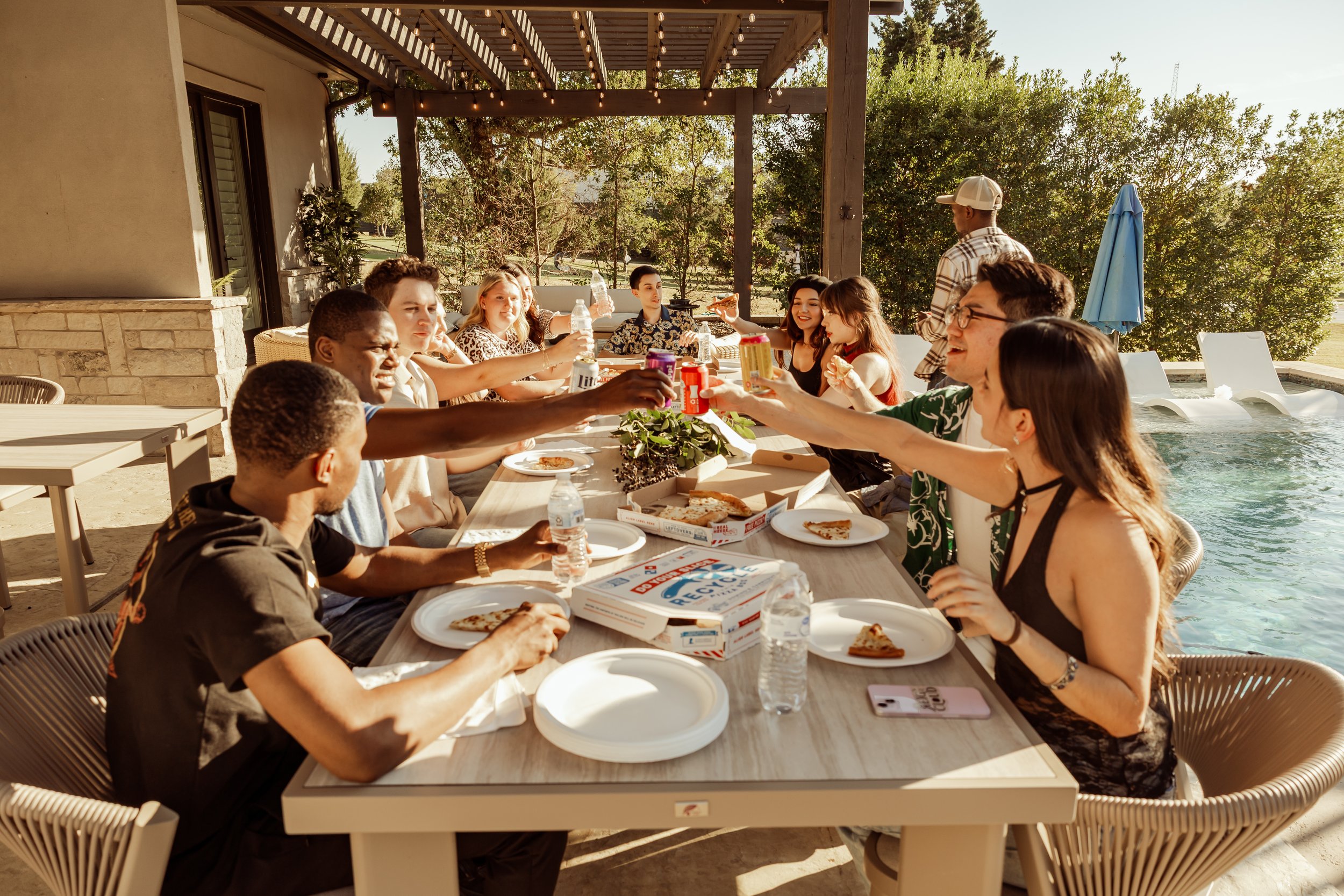 Group of people gathered around a long table outdoors by a pool, enjoying pizza and drinks, raising their glasses in a toast.