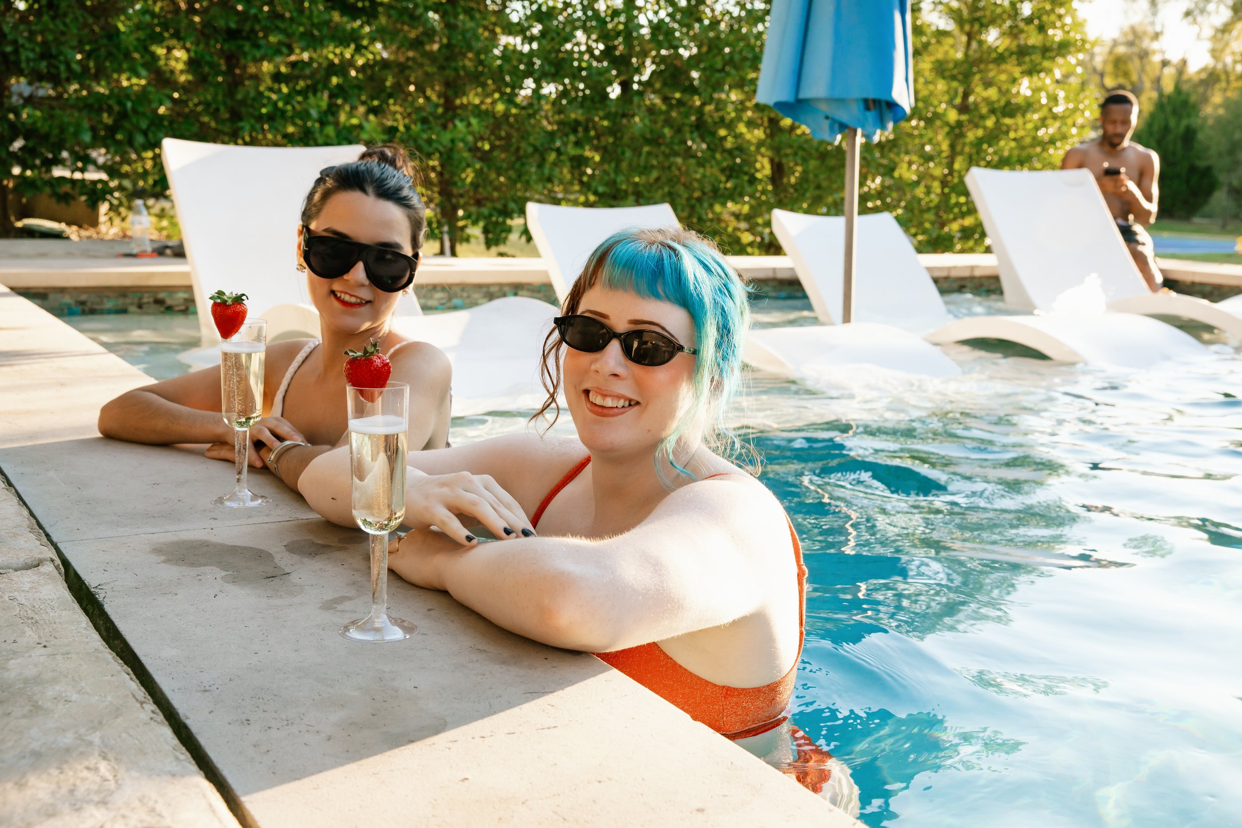Two women with colorful hair and sunglasses relaxing in a pool with strawberries and champagne, with a man in the background using his phone.