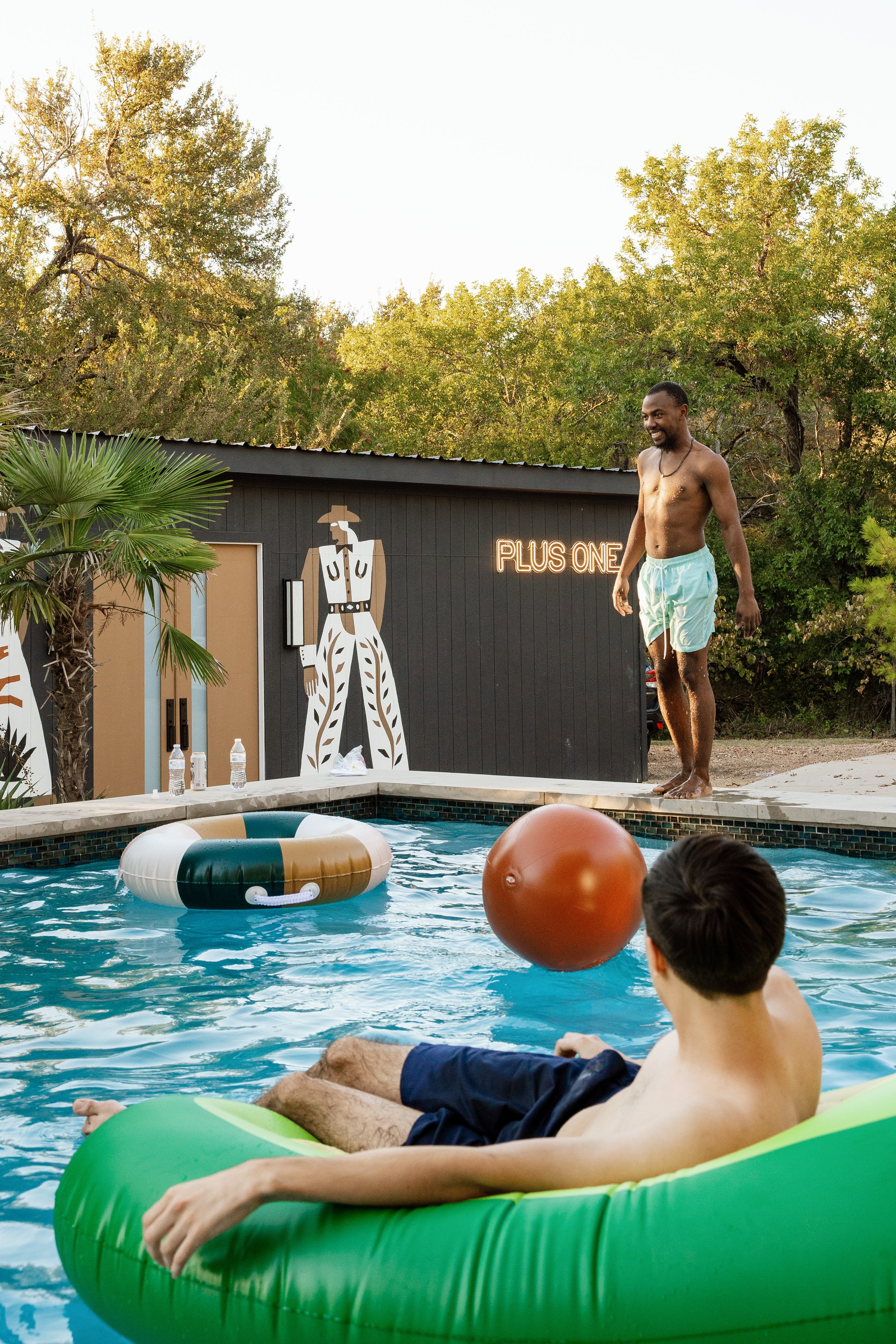 Two young men enjoying a swim in a backyard pool with a black building and green trees in the background. One man is sitting on a green inflatable tube in the pool, while the other man is standing at the edge of the pool, preparing to jump in. The po