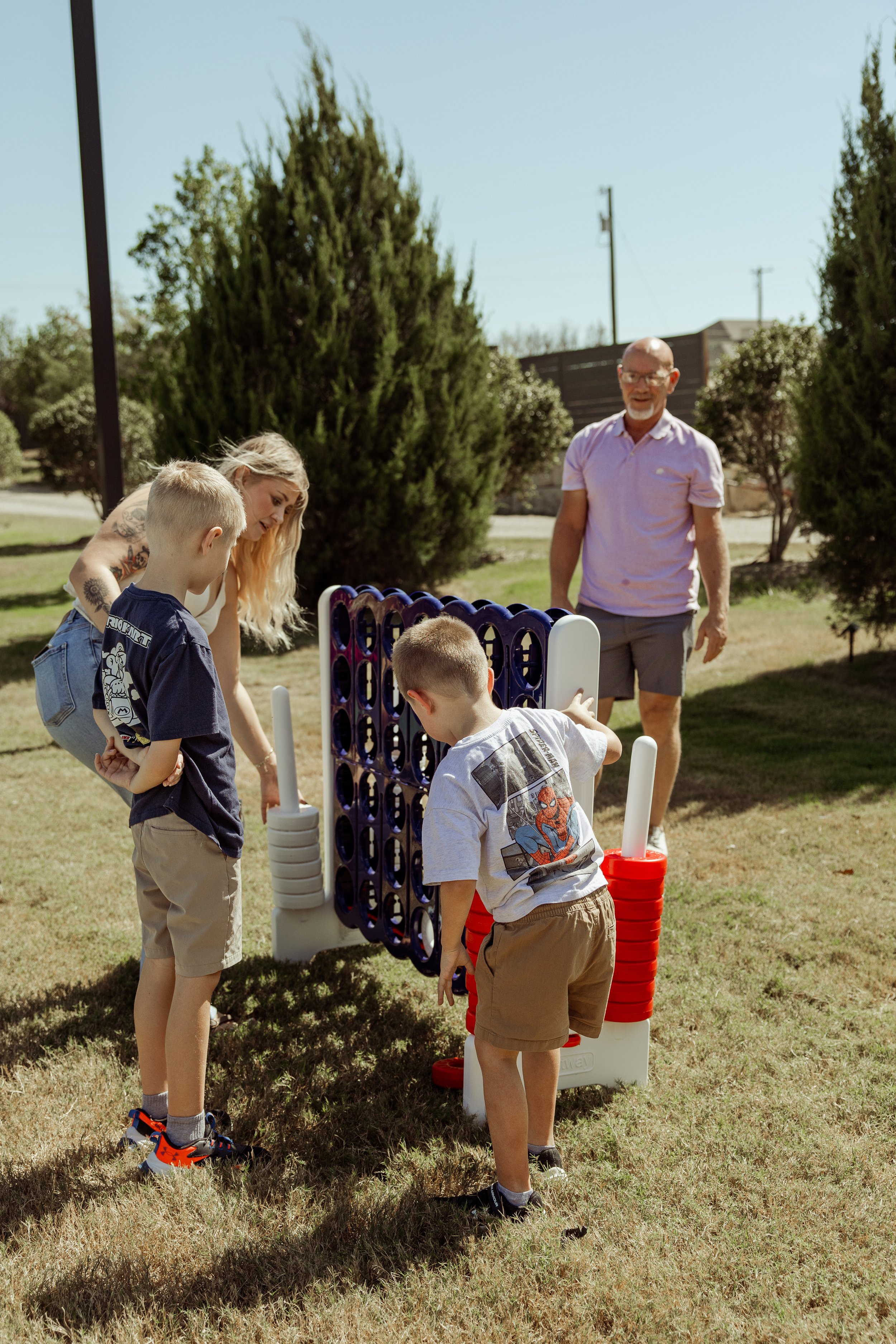 People playing giant Connect Four game outdoors on a sunny day, surrounded by trees.
