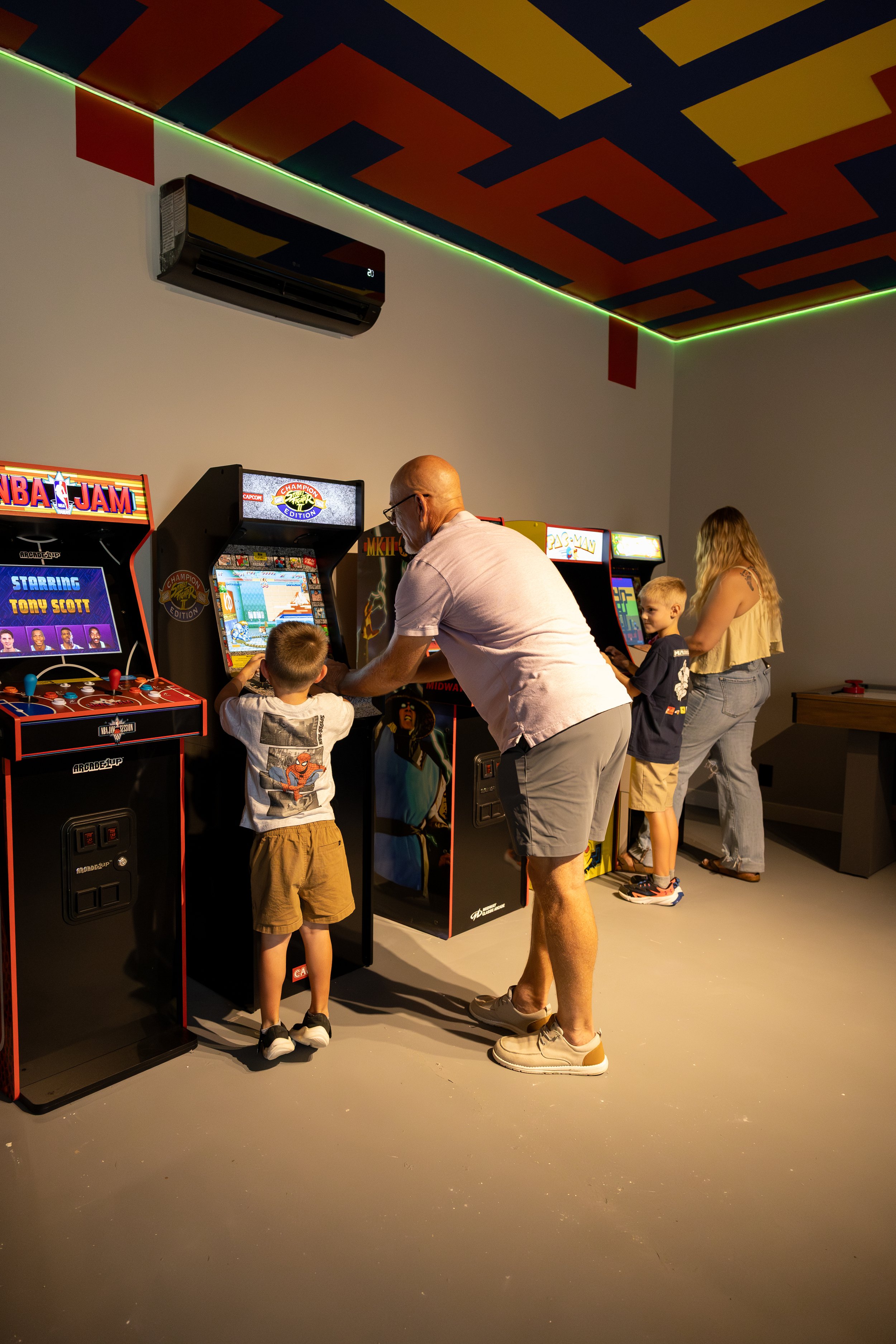 An adult and three children playing arcade video games in an arcade room with colorful ceiling decorations.