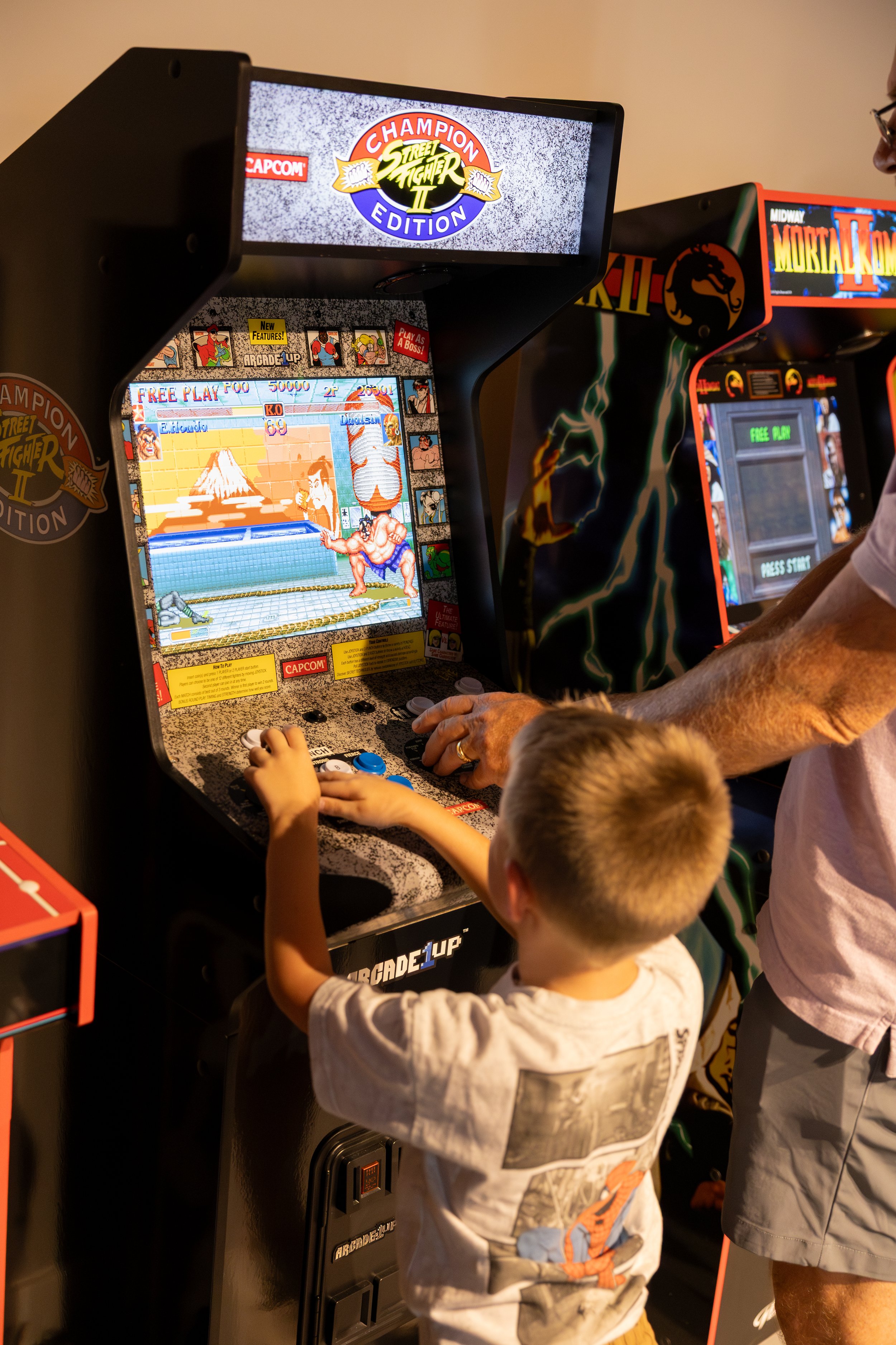 A young boy and an adult playing Street Fighter II arcade game in an arcade room, with another arcade game in the background.