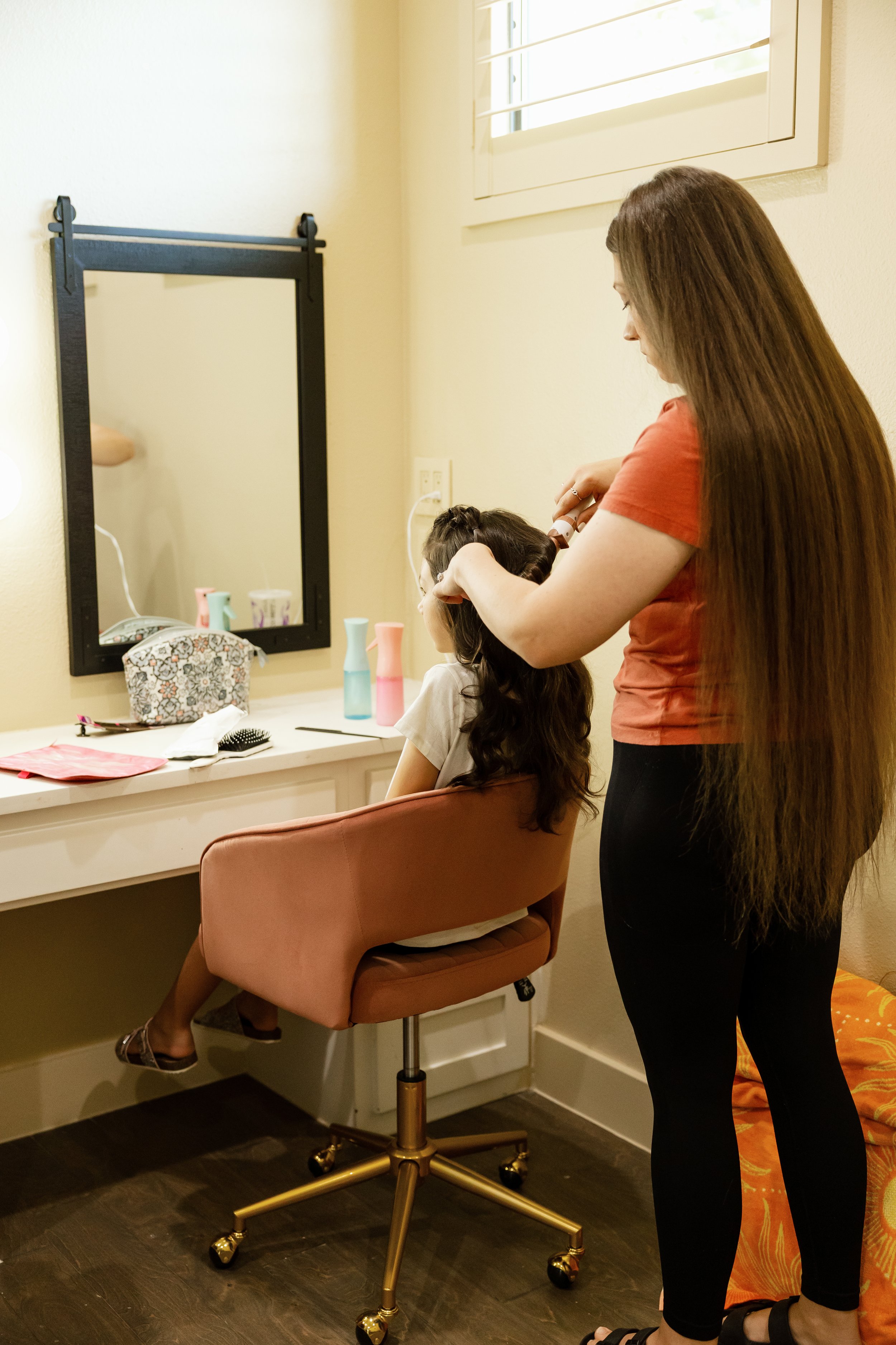 A woman with long brown hair wearing a red shirt and black pants is curling a young girl's hair in a dressing room. The girl is sitting on a pink chair and looking into a mirror while the woman styles her hair. The room has a white vanity with various hair styling tools and a window with blinds.