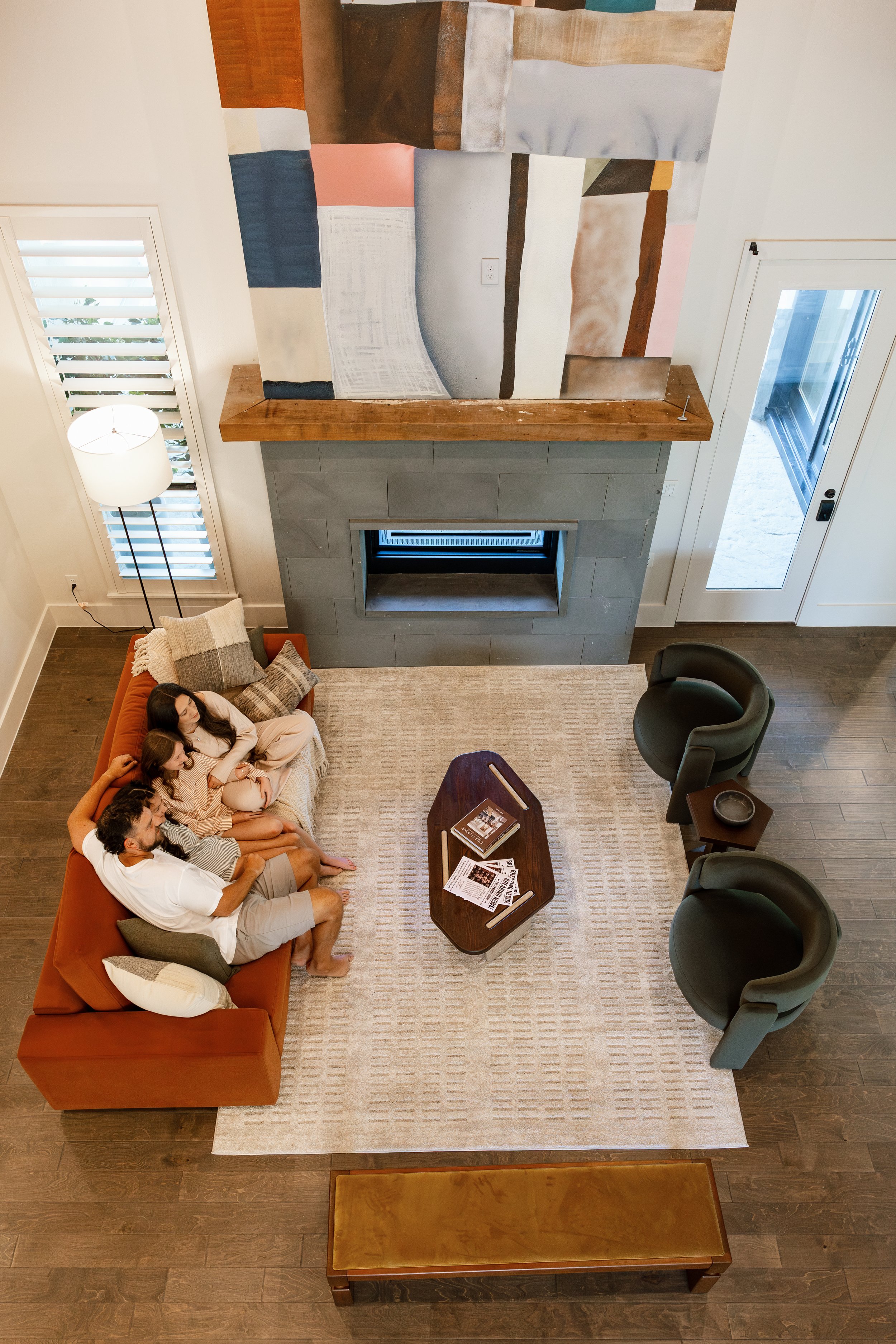 Living room with a fireplace, two green chairs, an orange sofa with four people, a coffee table with magazines, a rug, a wooden bench, a floor lamp, a sliding glass door, and a large abstract painting above the fireplace.
