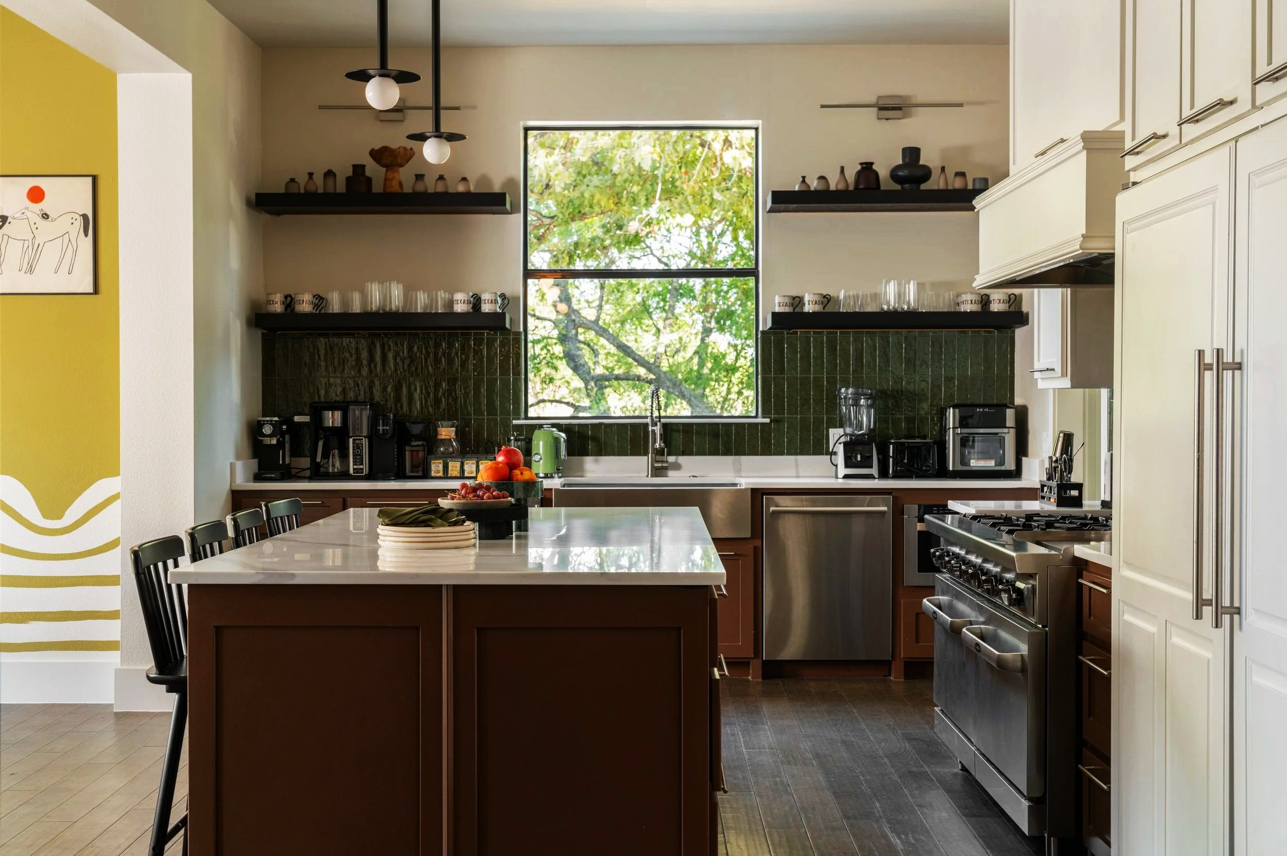 Modern kitchen with dark green tile backsplash, white upper cabinets, a brown island, and a window showing green trees outside.
