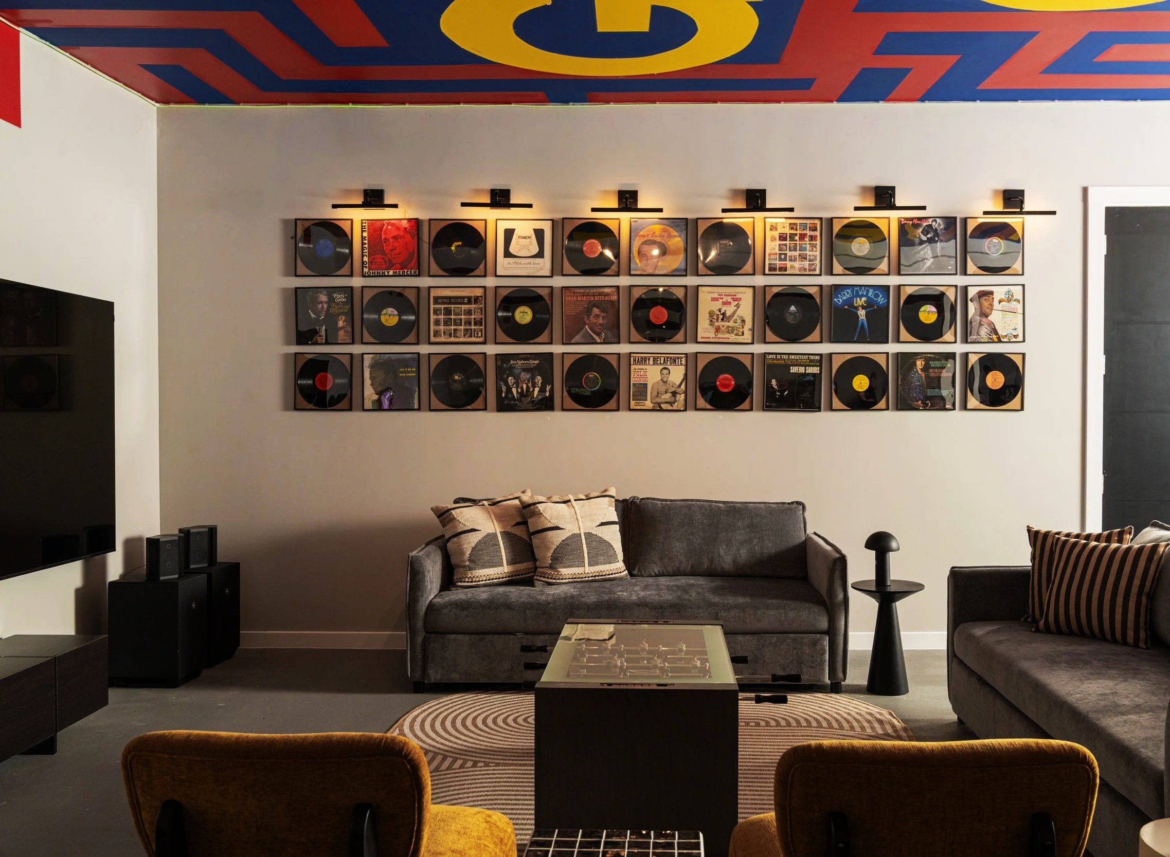 A cozy living room with a white wall decorated with framed vinyl records and album covers, two gray sofas with patterned pillows, a glass-topped coffee table, a black side table, a TV on the left, and a gold-colored chair in the foreground.