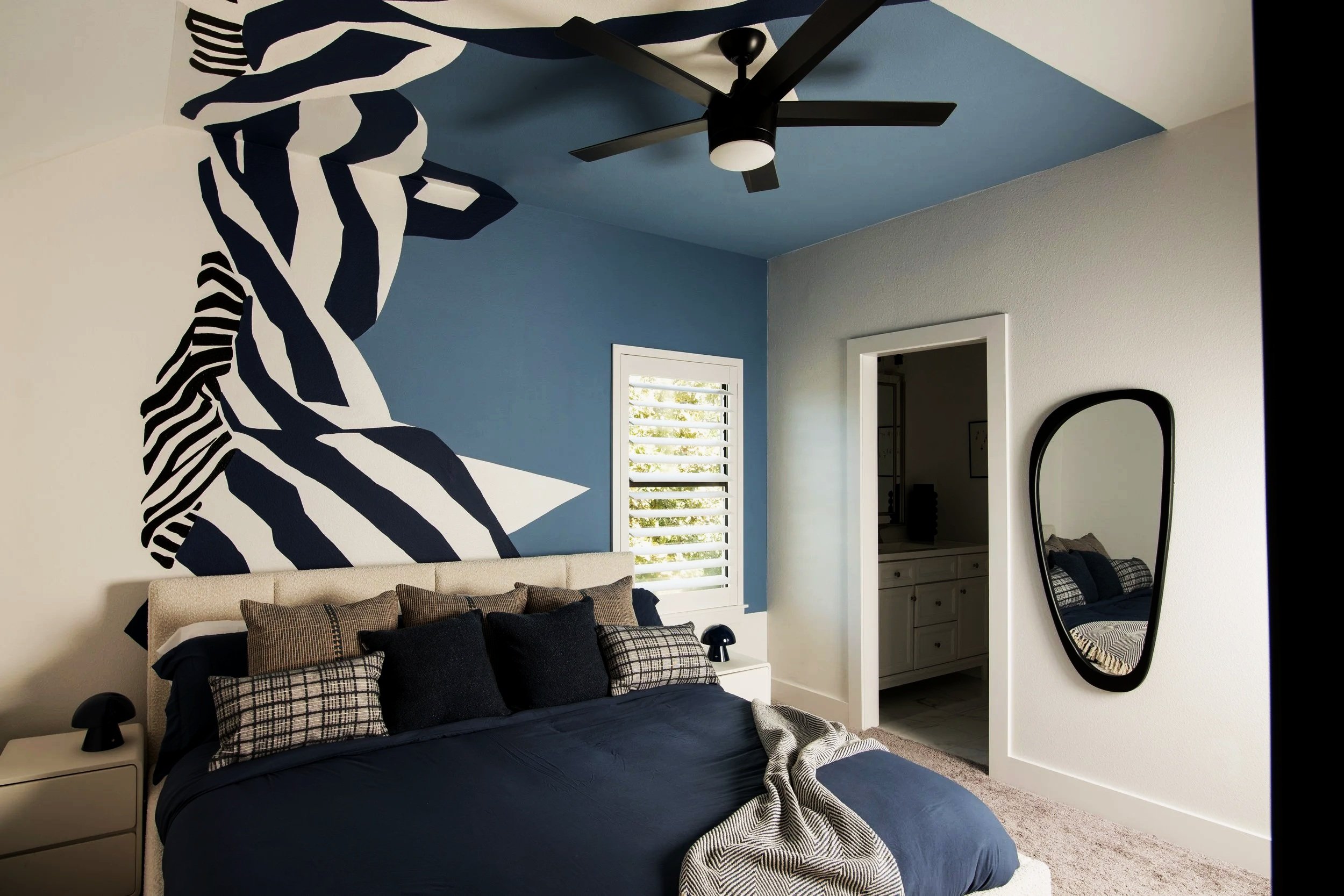 Bright bedroom with a beige upholstered bed against a wall with abstract navy and white mural art. The bed has various black, gray, and plaid pillows. To the right, a black-framed mirror and a window with blinds are visible. There's an open door lead
