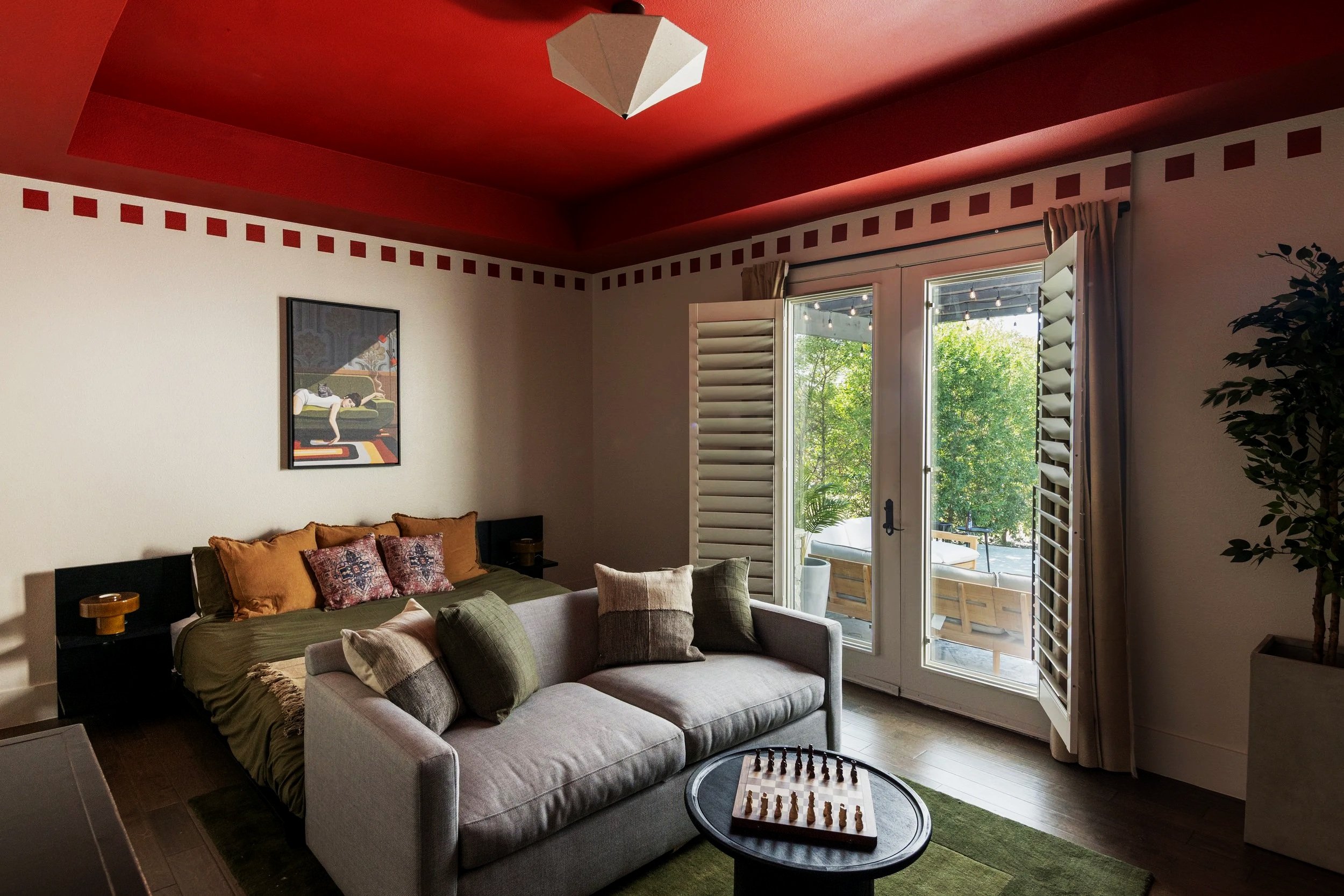 Living room with a gray sofa, decorative pillows, a chessboard, and a bed with pillows in the background, adjacent to a balcony with outdoor seating and greenery.