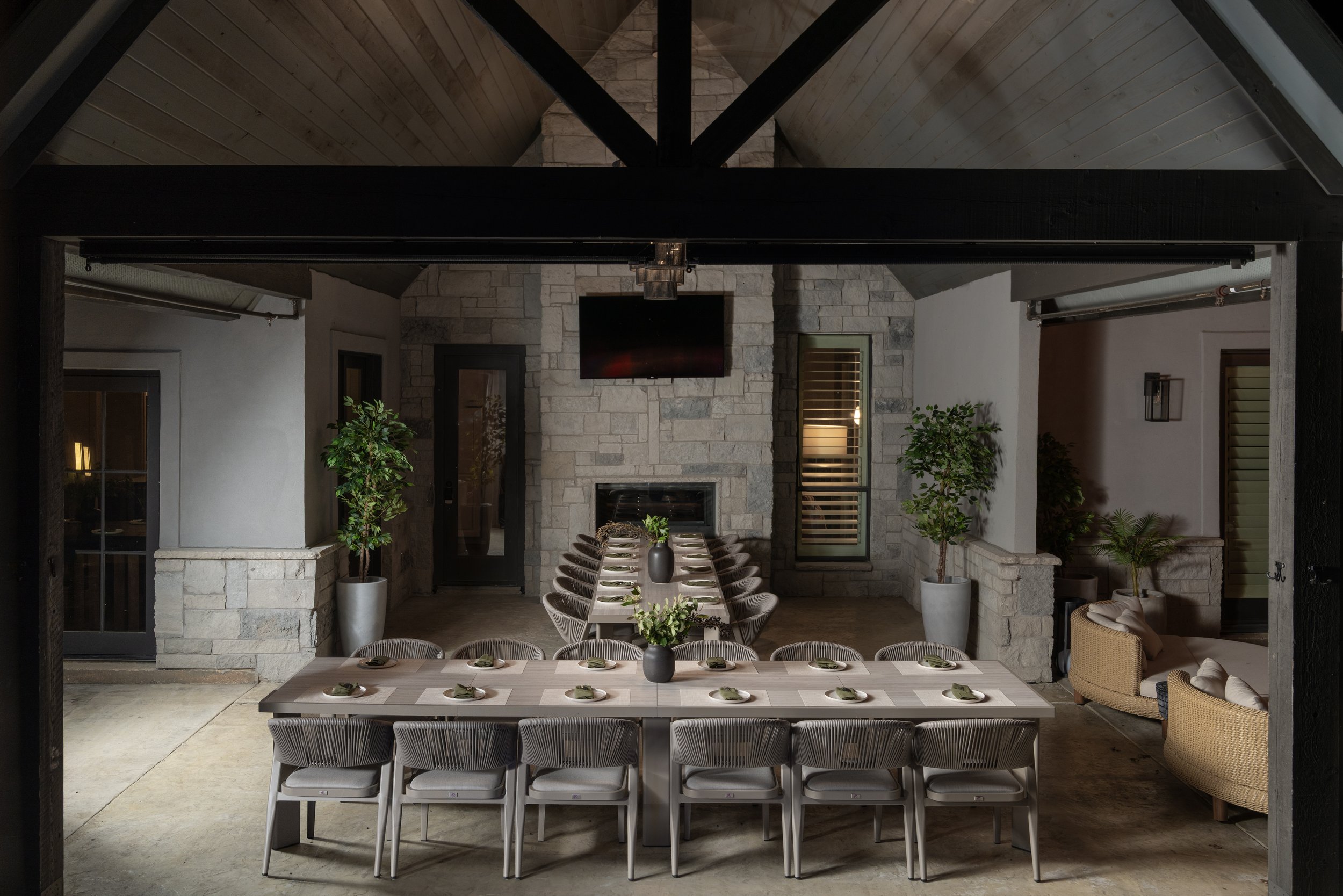Indoor dining area with two long tables set with plates and napkins, surrounded by chairs. The room has stone and white walls, large potted plants, and a stone fireplace with a mounted television above. There is a cozy seating corner with armchairs a