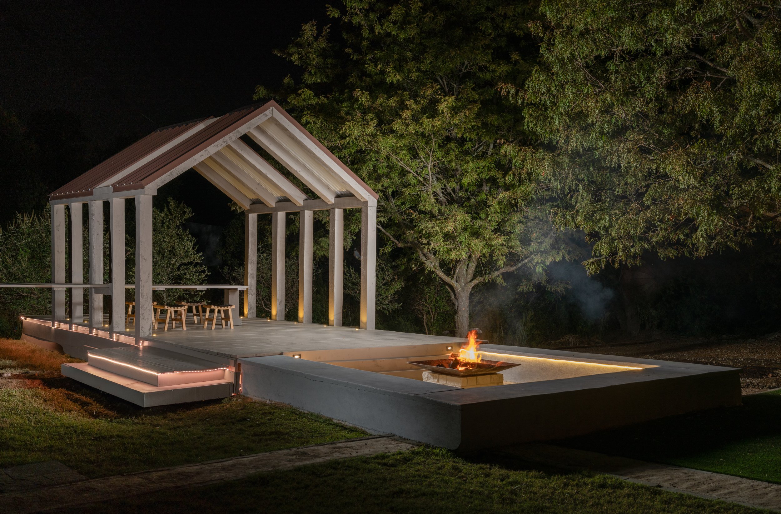 Nighttime outdoor patio with a fire pit, wooden seating, and a modern open-sided structure with a pitched roof, surrounded by trees.