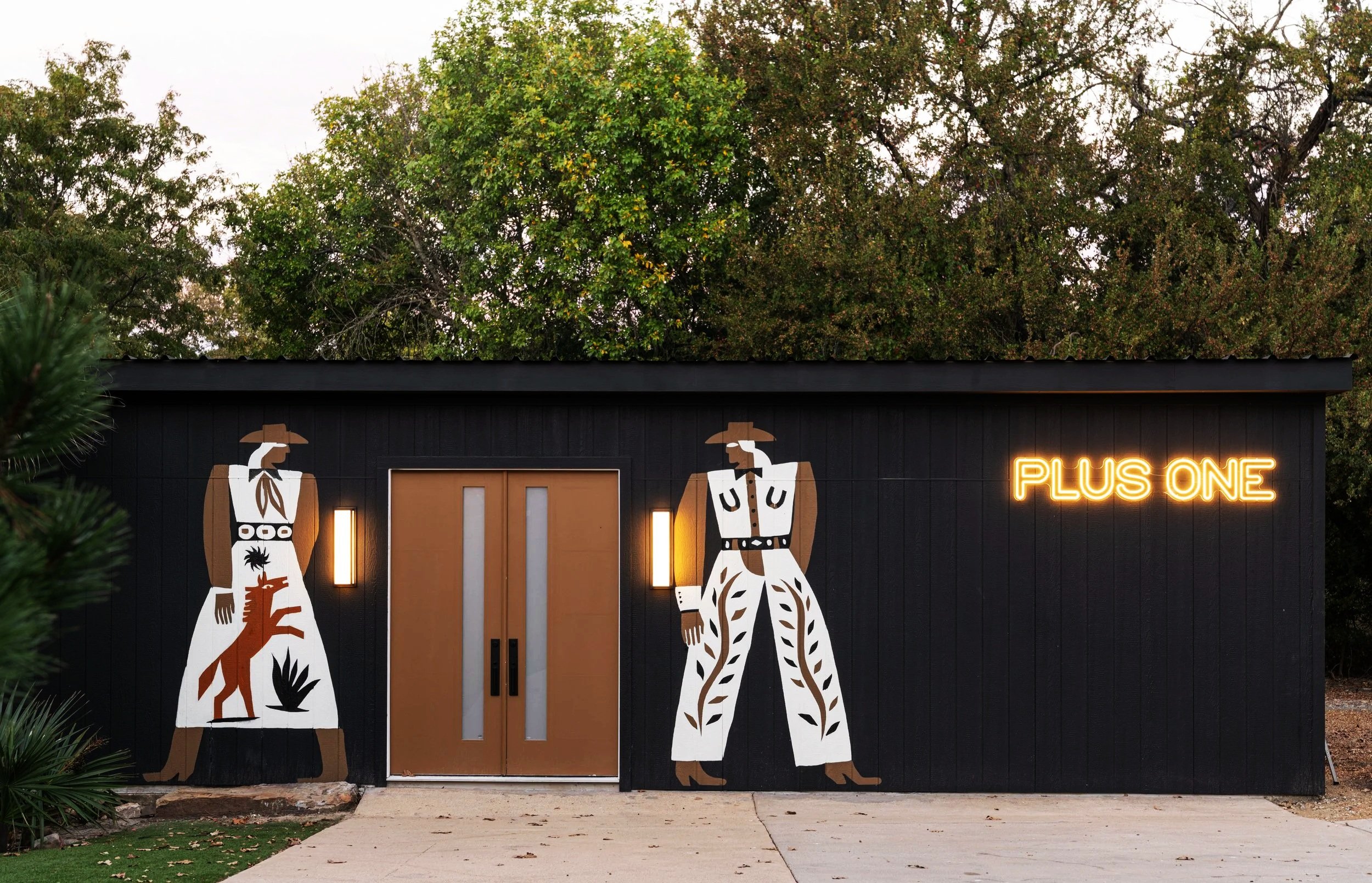 Exterior of a black building with two large cowboy-themed murals, a neon sign reading 'PLUS ONE,' and a brown door.