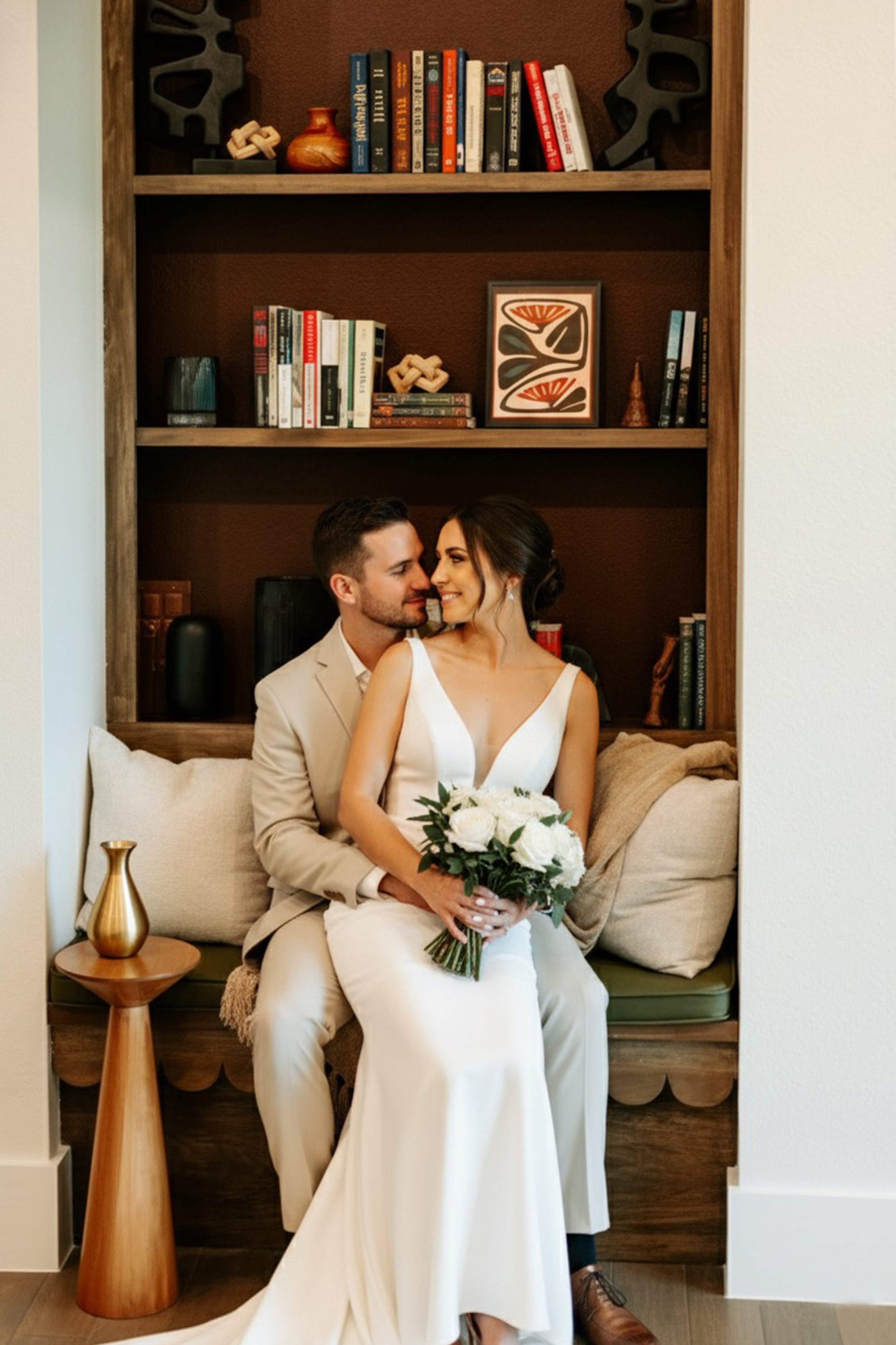 A newlywed couple sitting on a built-in bench with pillows, celebrating their wedding day. The bride is holding a bouquet of white flowers, and they're smiling at each other. The background features a wooden bookshelf with books and decorative items.