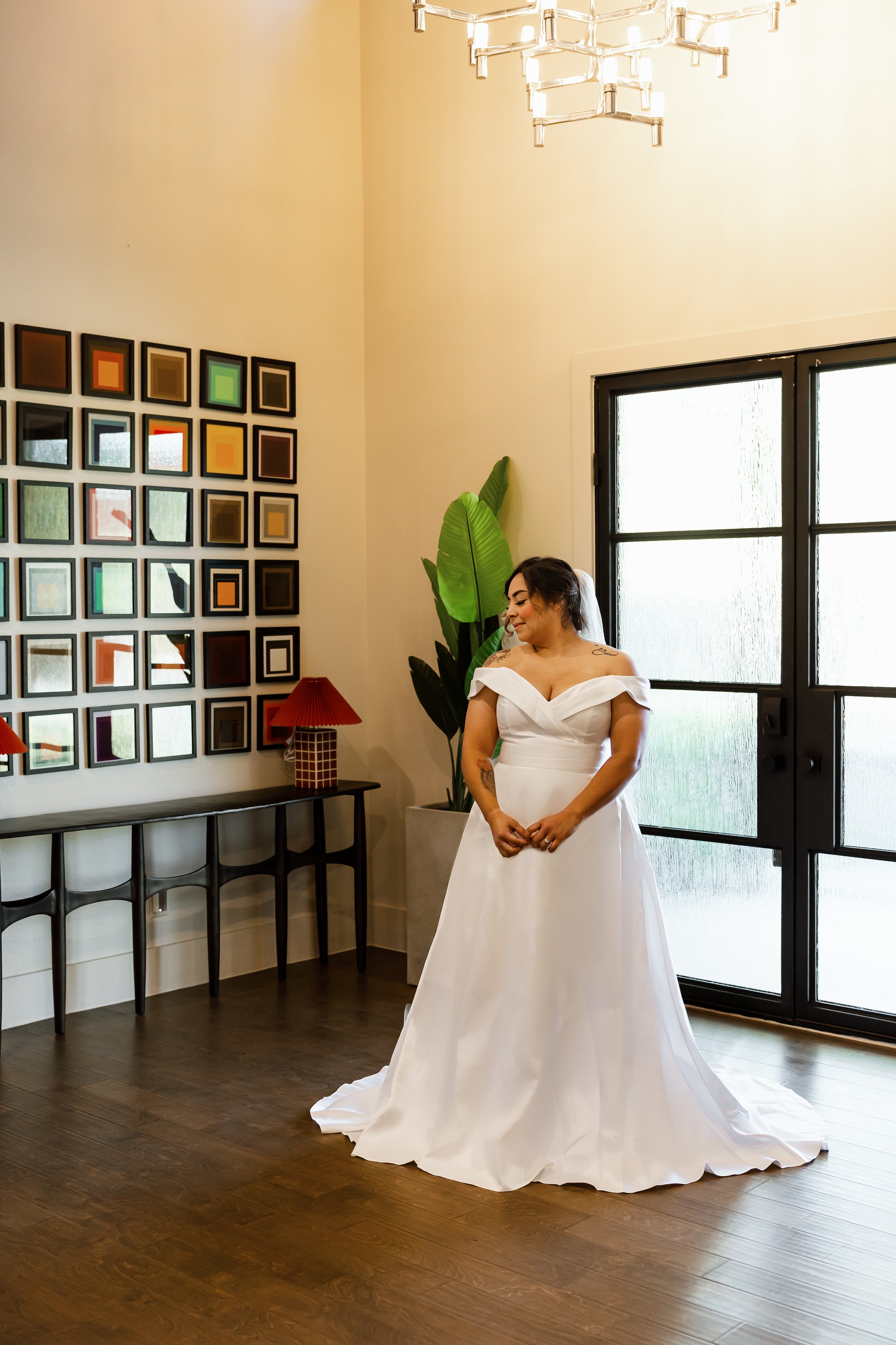 A woman in a white wedding dress standing indoors near a window, smiling with her hands clasped in front of her. There is a framed geometric art piece on the wall, a side table with two red lamps, a tall green plant, and wooden flooring.