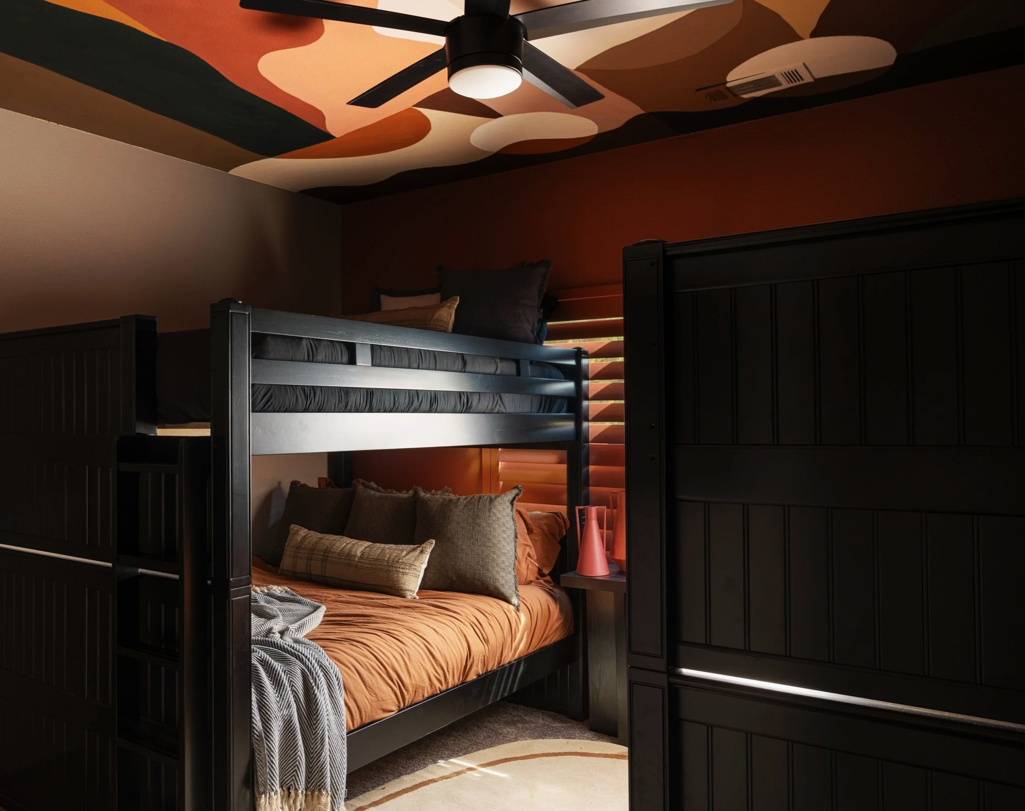 A bedroom with a black bunk bed, with an upper and lower bed, featuring dark linens and pillows. The room has a geometric pattern ceiling with warm colors, a ceiling fan, and a dark accent wall with a wooden headboard and a bedside lamp.
