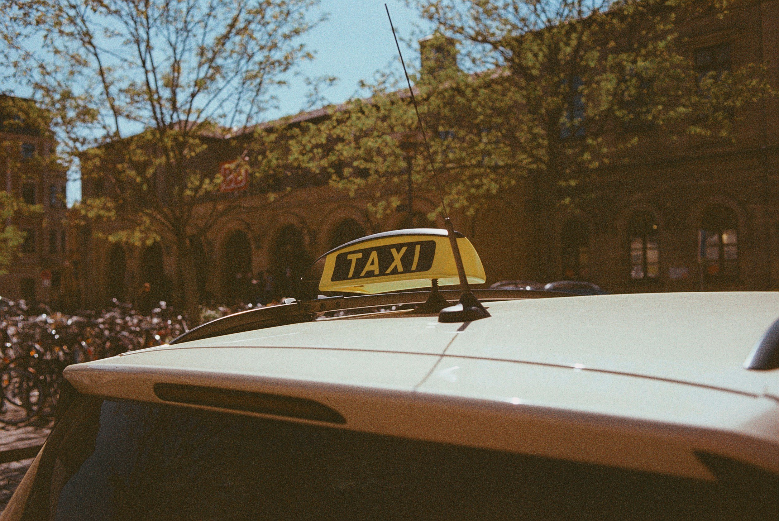 Close-up of a white taxi car with a yellow taxi sign on top, parked outside on a city street with trees and old-style buildings in the background.