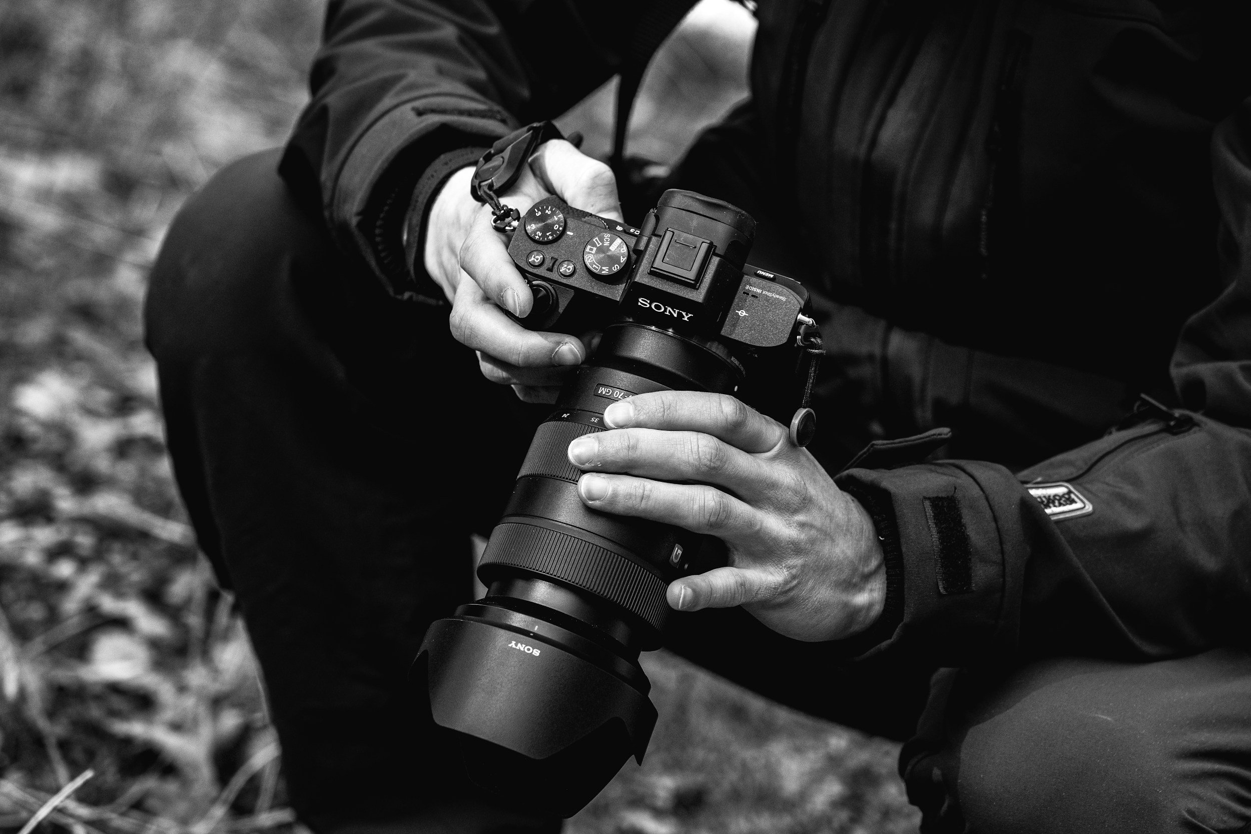 A person holding a Sony mirrorless camera with a large telephoto lens attached, outdoors on a grassy surface.