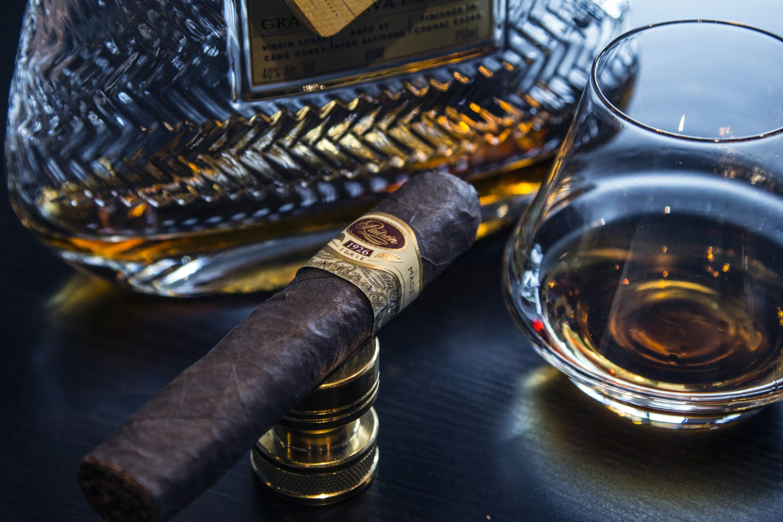 A cigar with a gold band resting on a gold ring, a glass of whiskey, a bottle of whiskey in the background, and a whiskey glass on a dark surface.