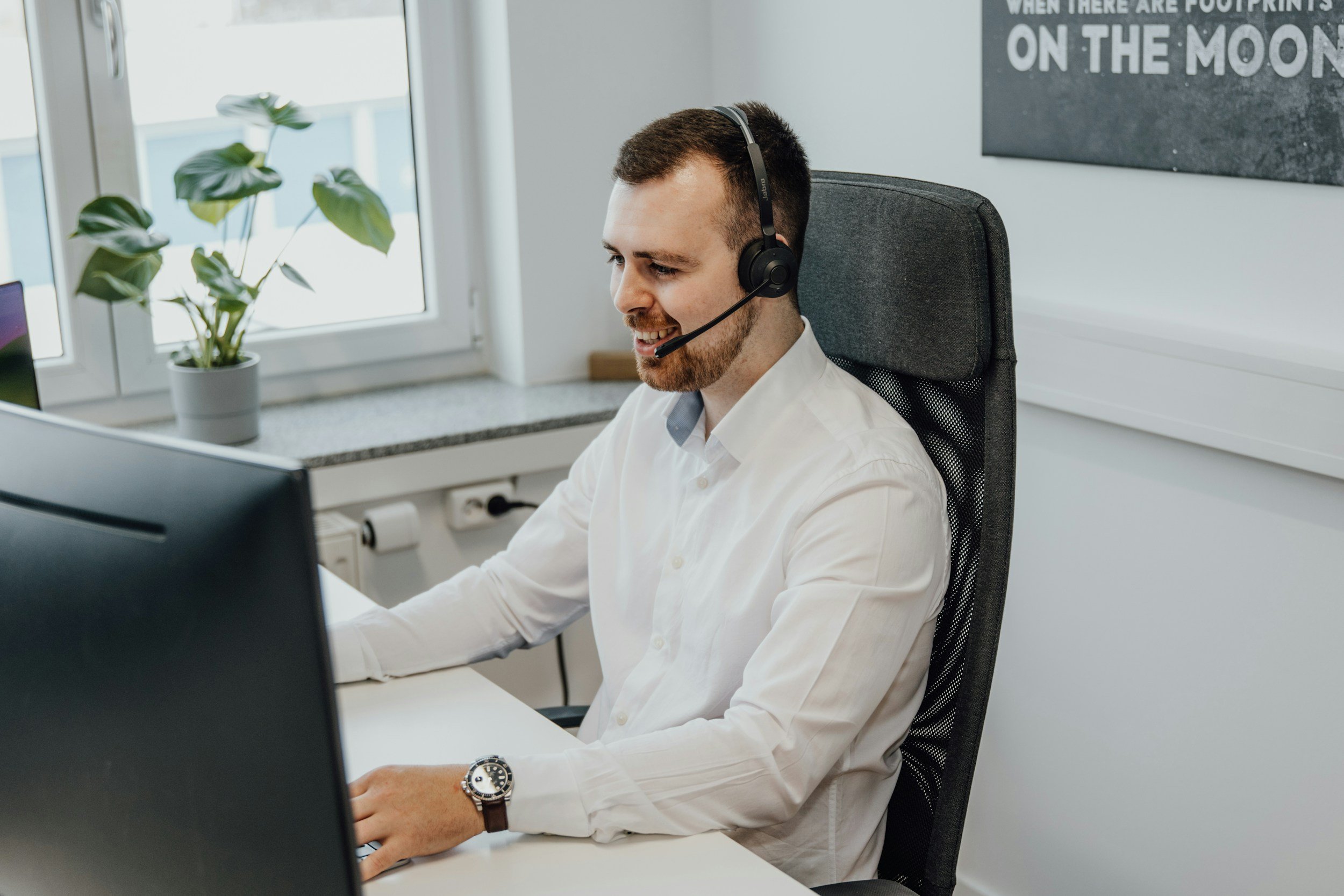 A man wearing a white shirt and a watch, sitting in an office chair at a desk, using a computer with a headset on, in a bright office with a window, a potted plant, and a poster on the wall.