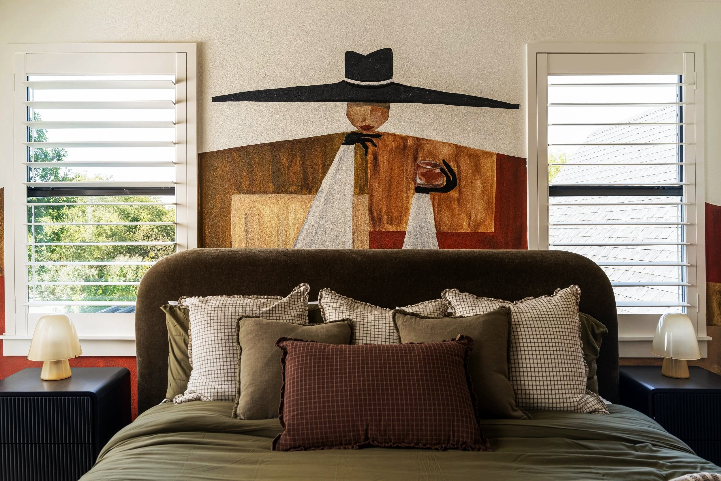 Decorative bedroom wall art depicting a stylized woman in a wide-brimmed hat, holding a glass, with two windows with white shutters on either side, and a bed with multiple pillows and dark headboard in front.