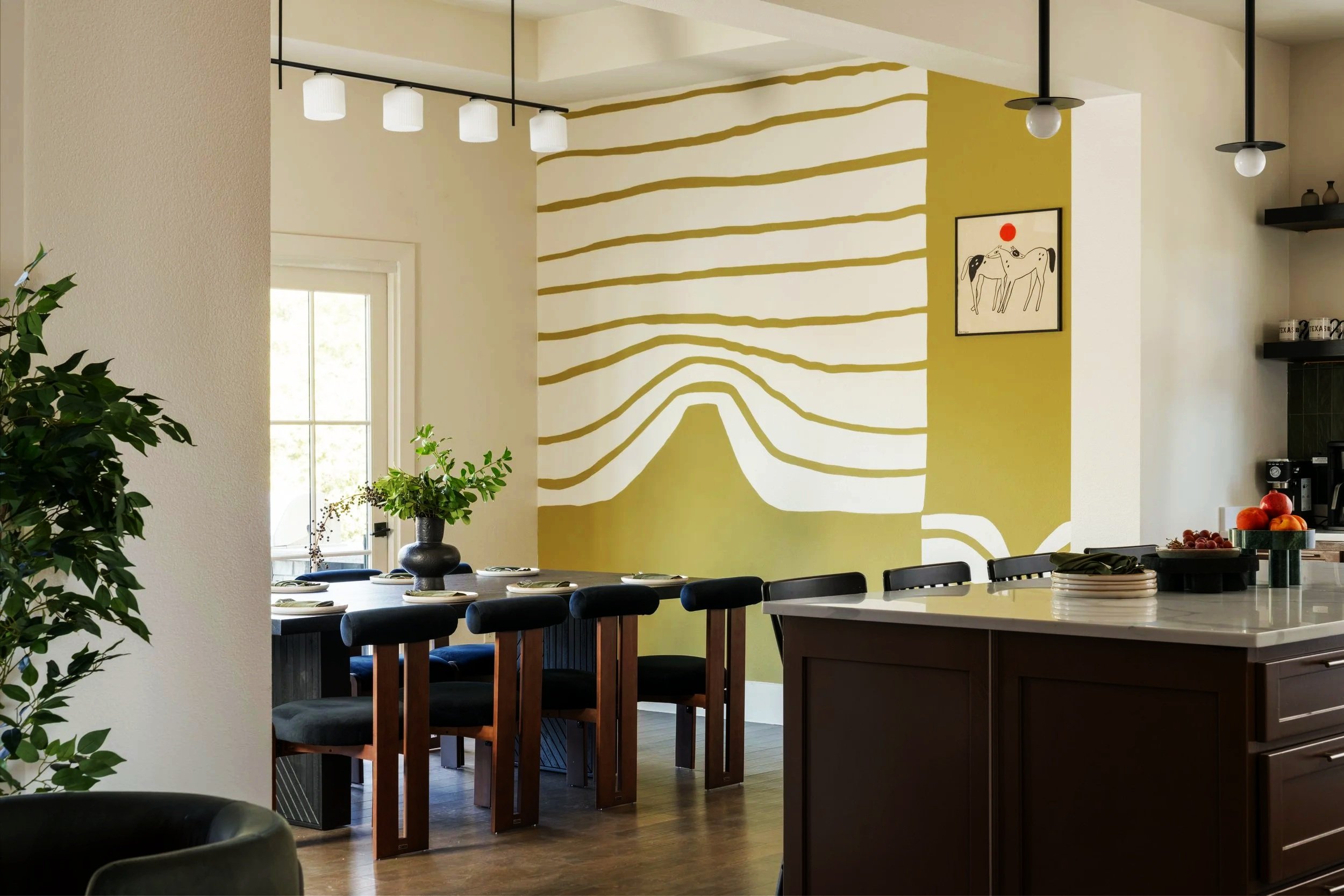 Modern dining area with a black table and chairs, a plant centerpiece, and a wall with abstract yellow and white striped mural, adjacent to a kitchen with dark cabinets and a white countertop.