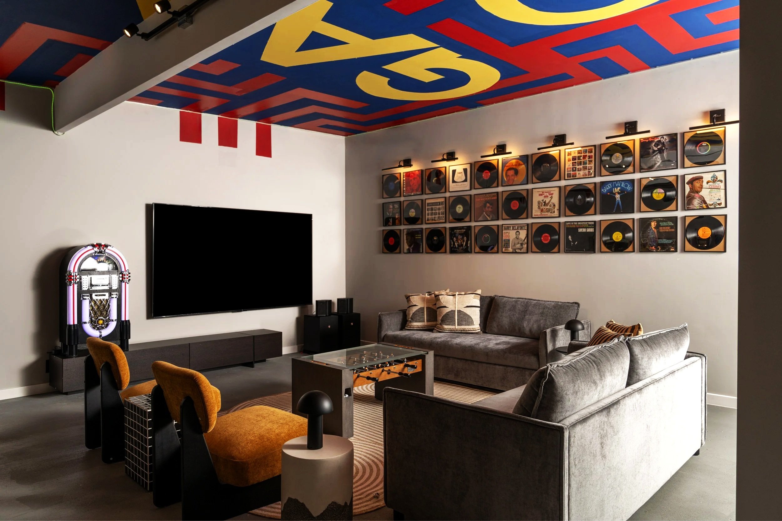 Modern living room with a large flat-screen TV, vintage jukebox, gray sofas, orange chairs, music album covers on the wall, and a colorful ceiling mural.