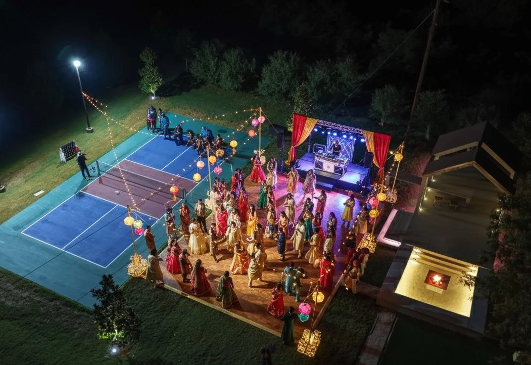 A nighttime outdoor celebration with people dancing on a wooden dance floor, decorated with hanging lanterns, next to a stage with a DJ mixer. There are tennis courts on the left and a small gathering of people on the court. The area is well-lit with string lights and surrounded by trees.