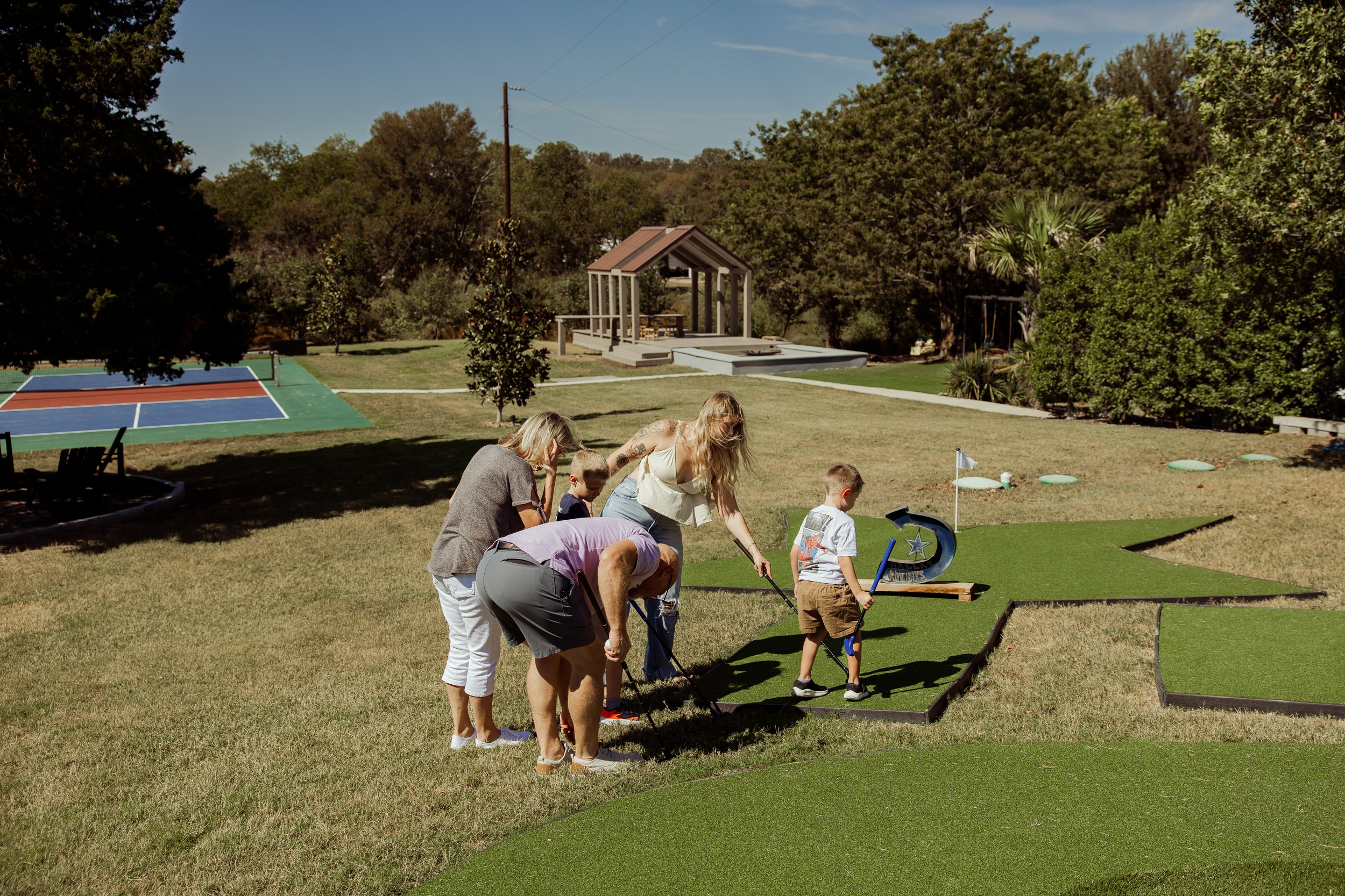 A group of people, including children and adults, playing mini-golf outdoors on artificial turf on a sunny day, with a park and trees in the background.