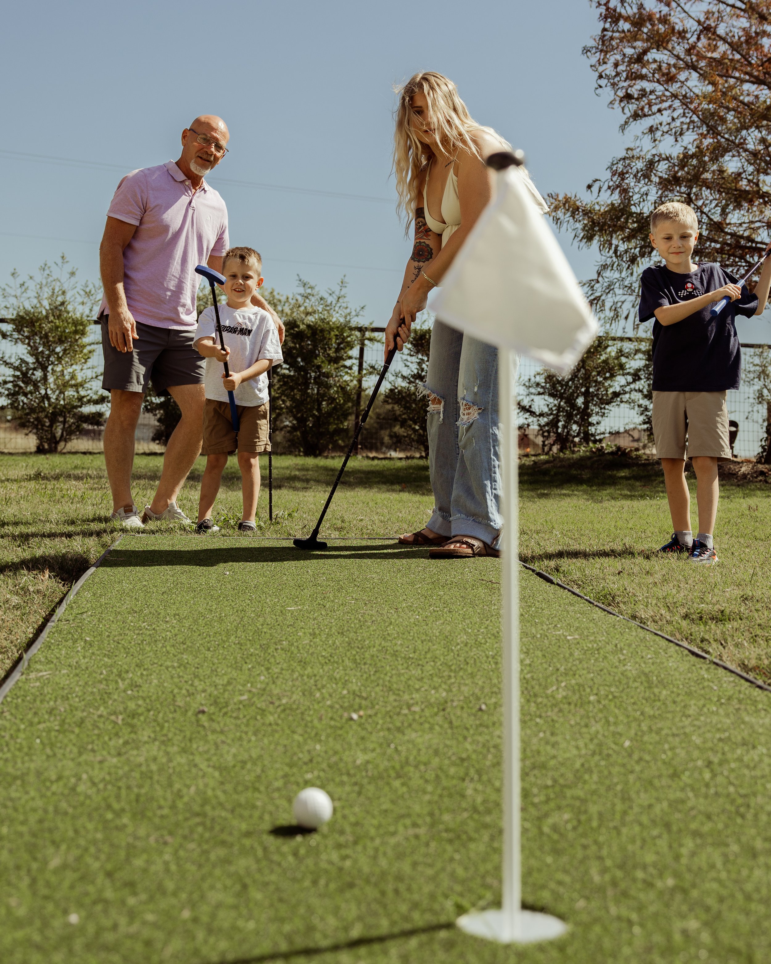 Family playing mini golf outdoors on a sunny day, with a green course, flag, and trees in the background.