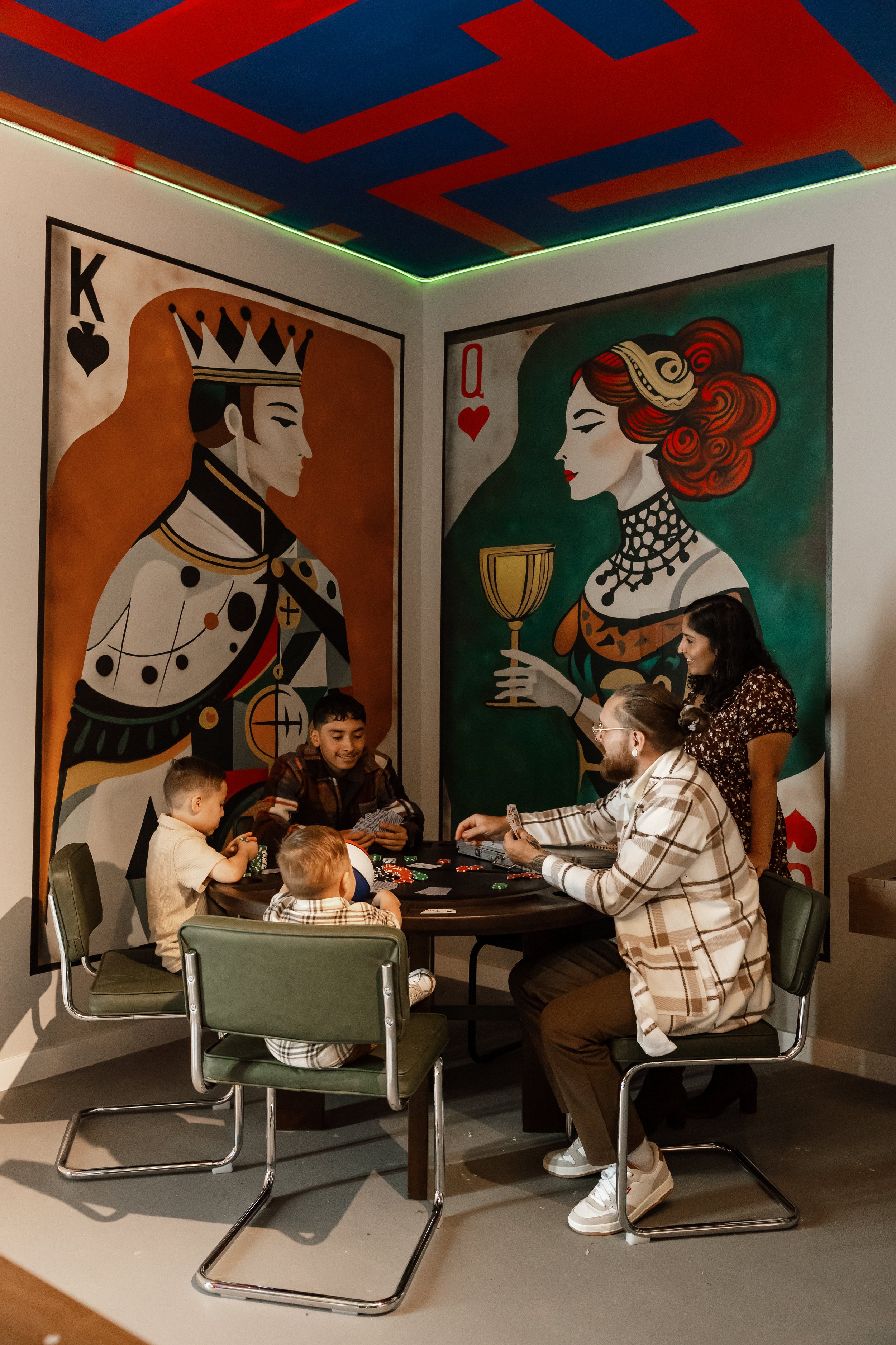A group of people, including children and adults, playing poker around a table in a room decorated with large playing card wall art.
