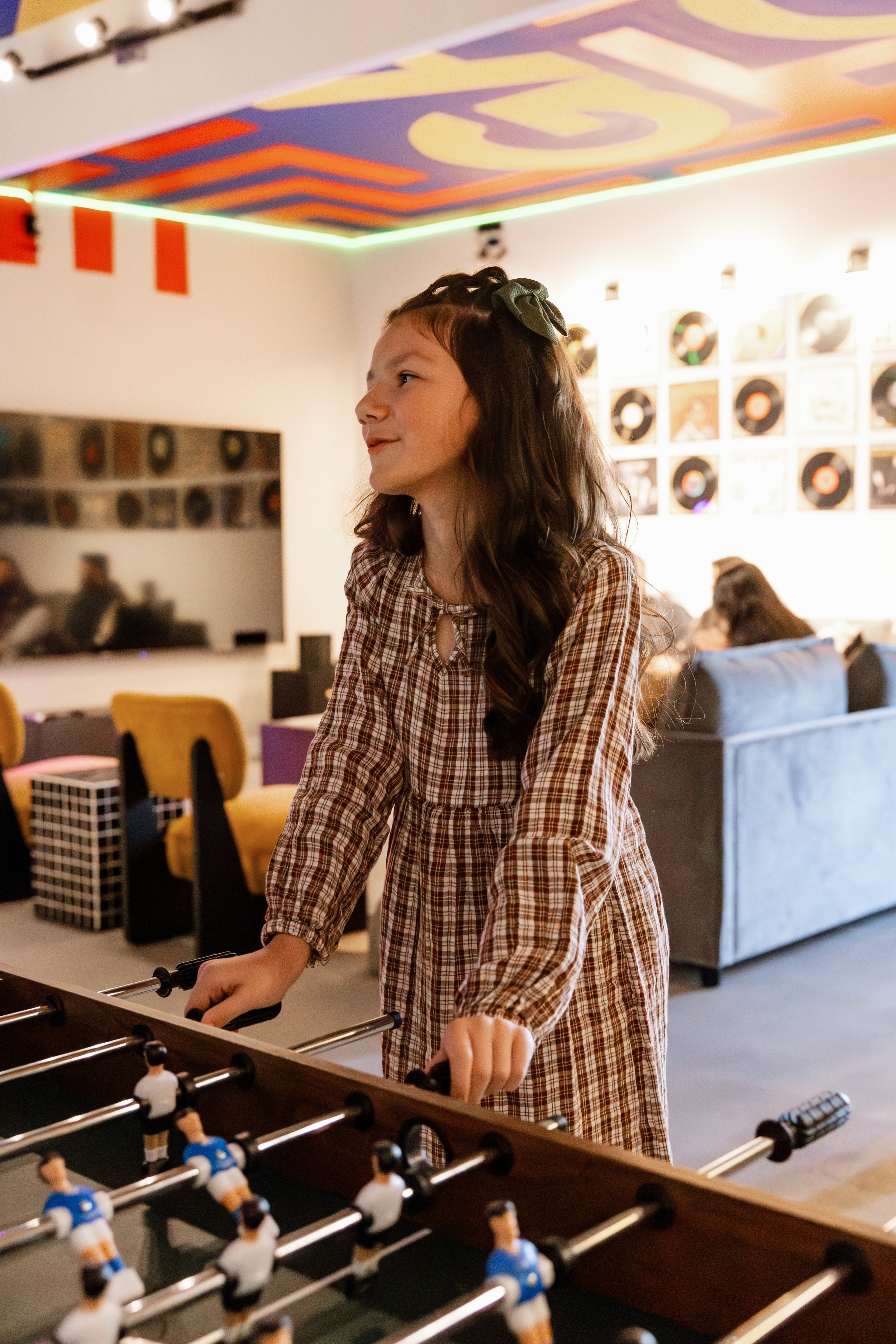 A young girl with long dark hair and a green bow, wearing a brown plaid dress, is playing foosball in a room decorated with colorful artwork and records.