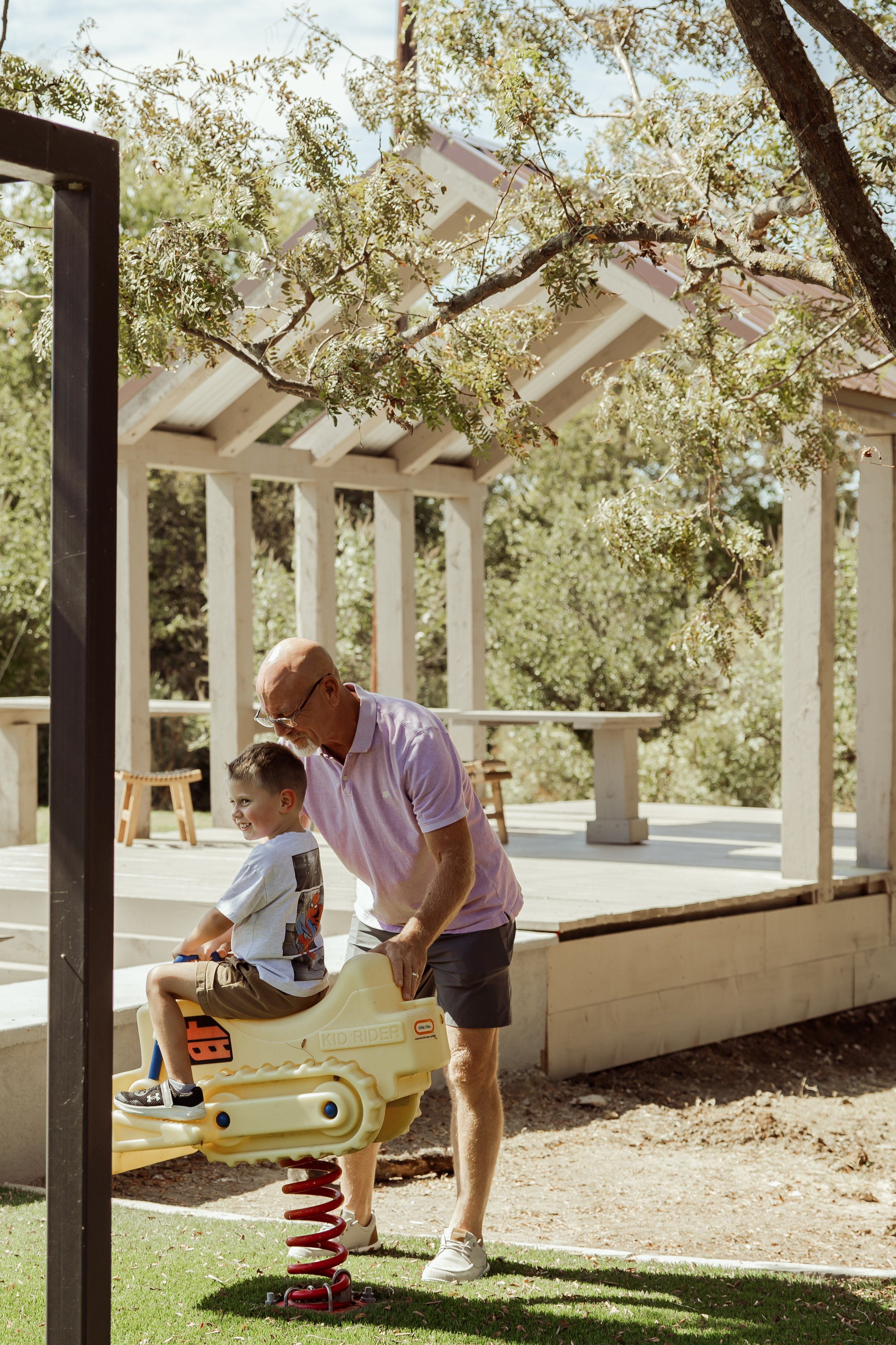An elderly man pushing a young boy on a spring rider toy in a backyard with a wooden deck and green trees.