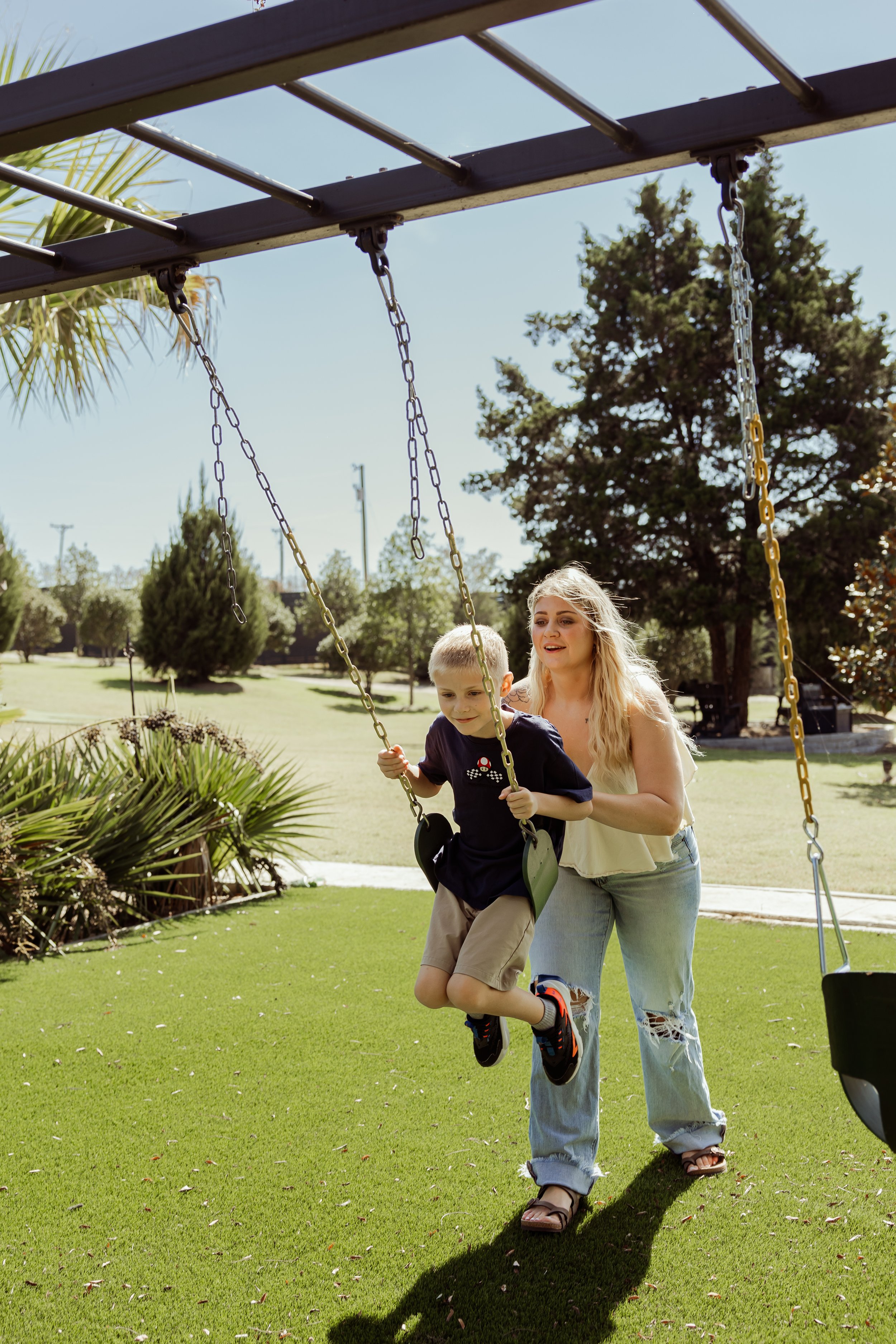 A woman helping a young boy on a swing set in a park on a sunny day.