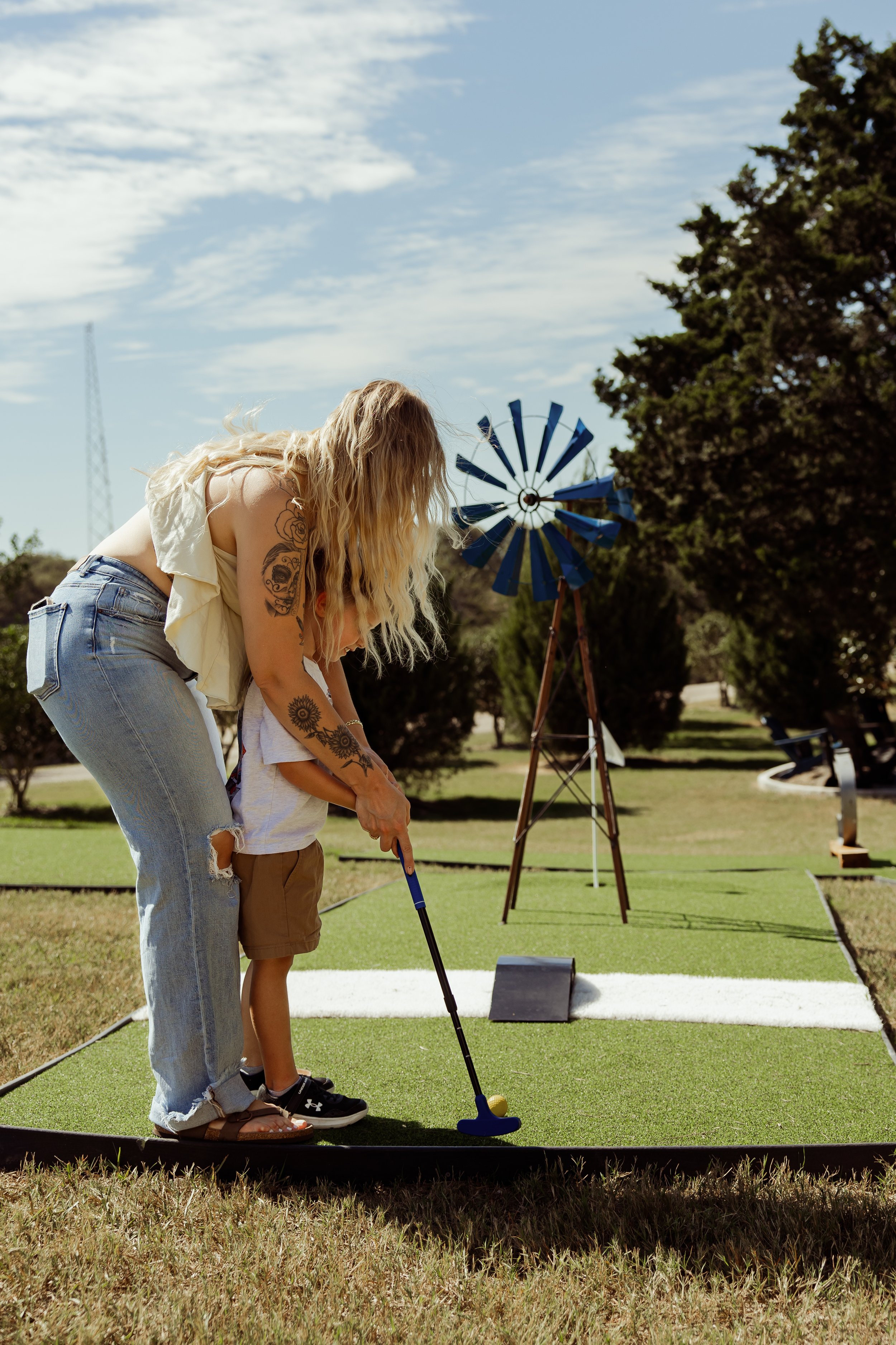 A woman with tattoos and long blonde hair teaching a young boy how to golf on a mini golf course outdoors, with a windmill and trees in the background.