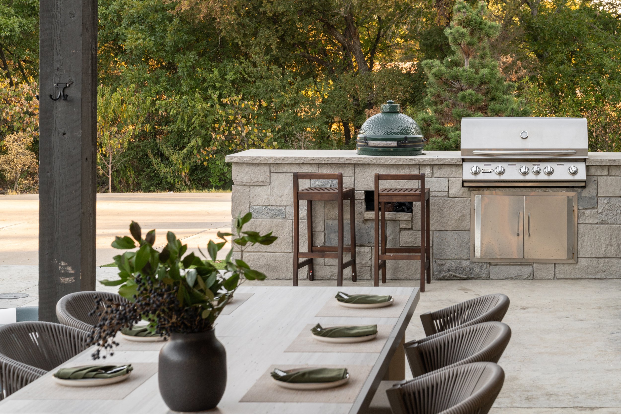 Outdoor patio dining area with a dining table set with four plates and napkins, a large vase with greenery, and a stone kitchen counter with a green grill and two wooden bar stools.