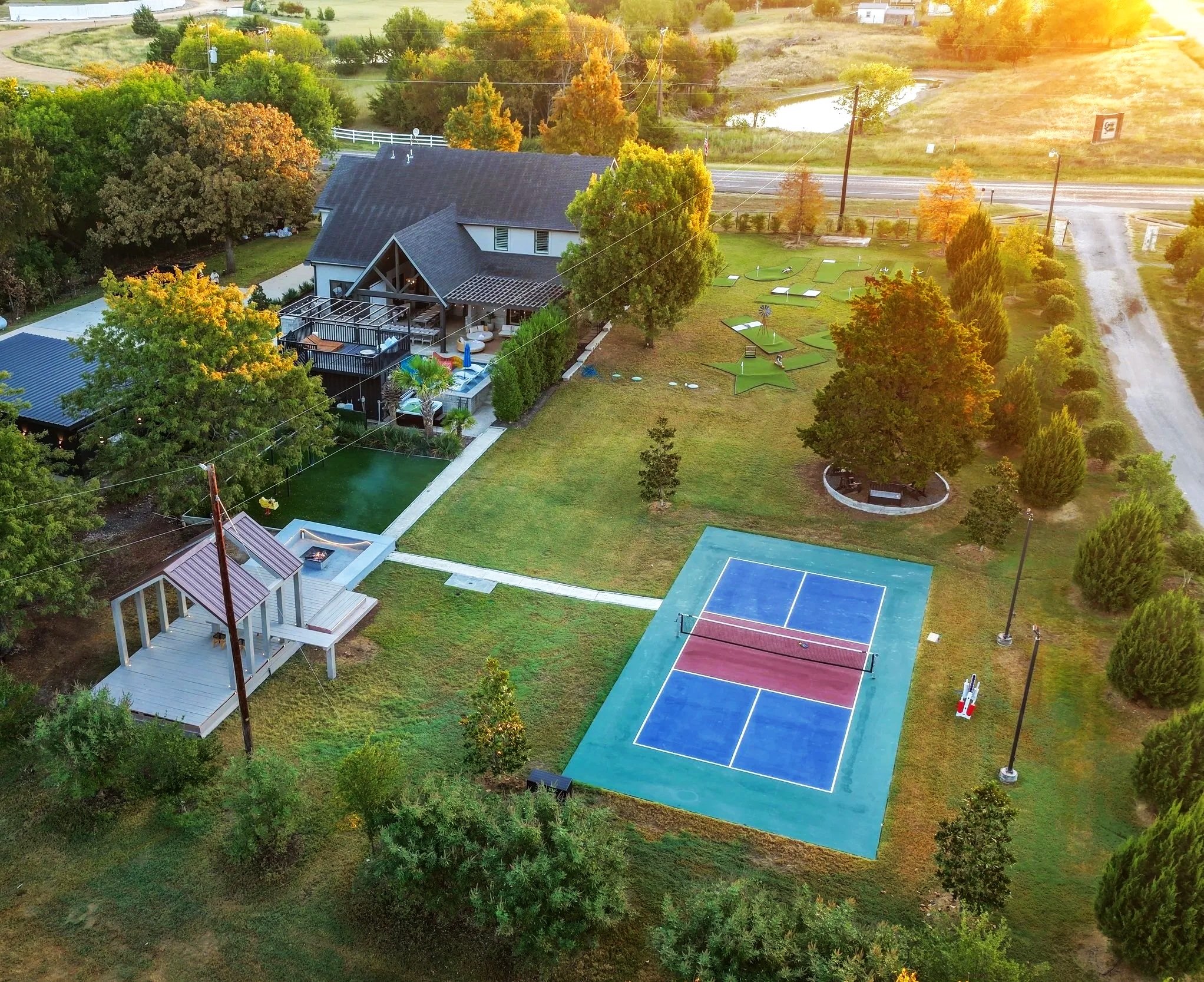 A backyard with a tennis court, a hot tub, a deck with string lights, trees, and decorative star-shaped lawn art, surrounded by a white fence and trees, with a road nearby and sunset lighting.