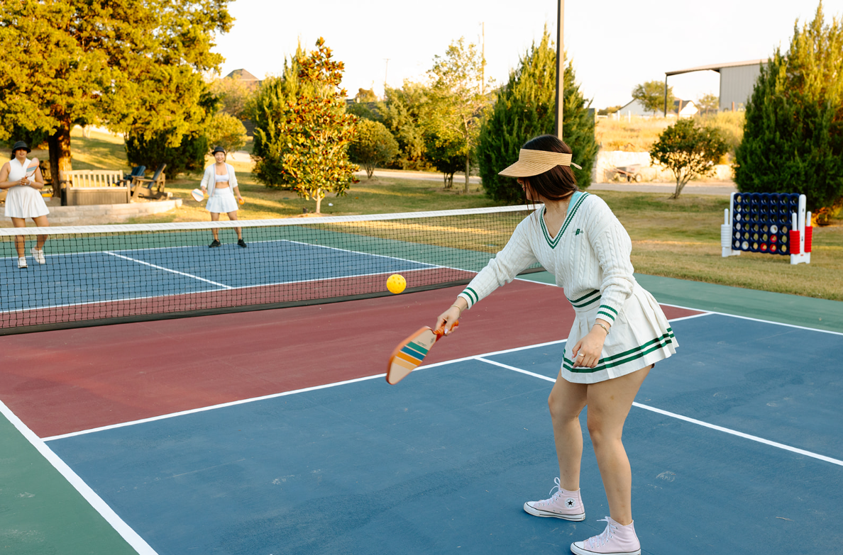 A woman playing pickleball on an outdoor court with trees and a person in the background.